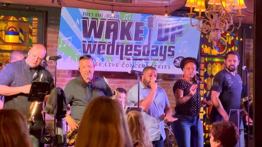 The Accents perform for a crowd on the Flynn Side at Ryan's Wake, with singers and a saxophonist in front of a Wake Up Wednesdays free live concert series banner and stained-glass windows.