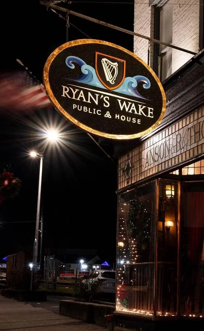 Nighttime image of a sign for Ryan's Wake Public & House with a harp emblem and blue wave design, illuminated and hanging outside a building.