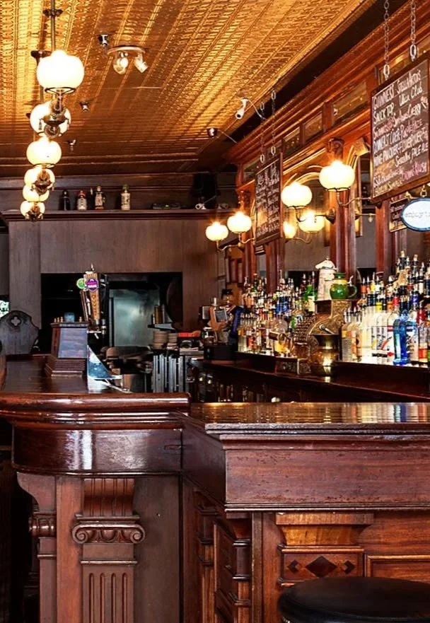 Interior of a wooden bar with illuminated hanging lamps, a variety of liquor bottles on the shelves, and chalkboard menus on the wall.