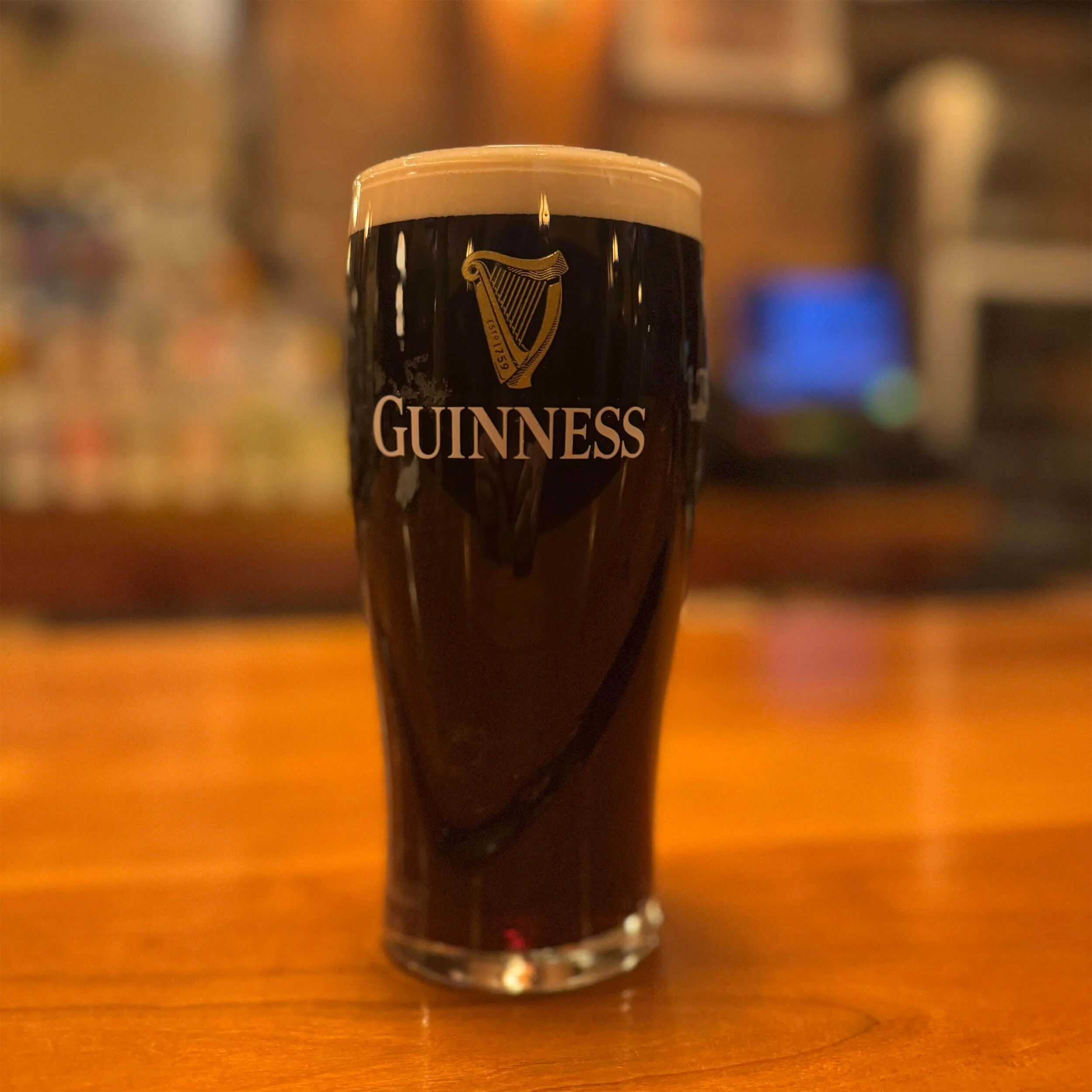 A pint glass filled with Guinness beer, topped with a creamy head, sitting on a wooden surface with a blurred background at Ryan's Wake | An Irish Pub, Bar & Restaurant in Troy, NY with River Views, Outdoor Seating & Live Music