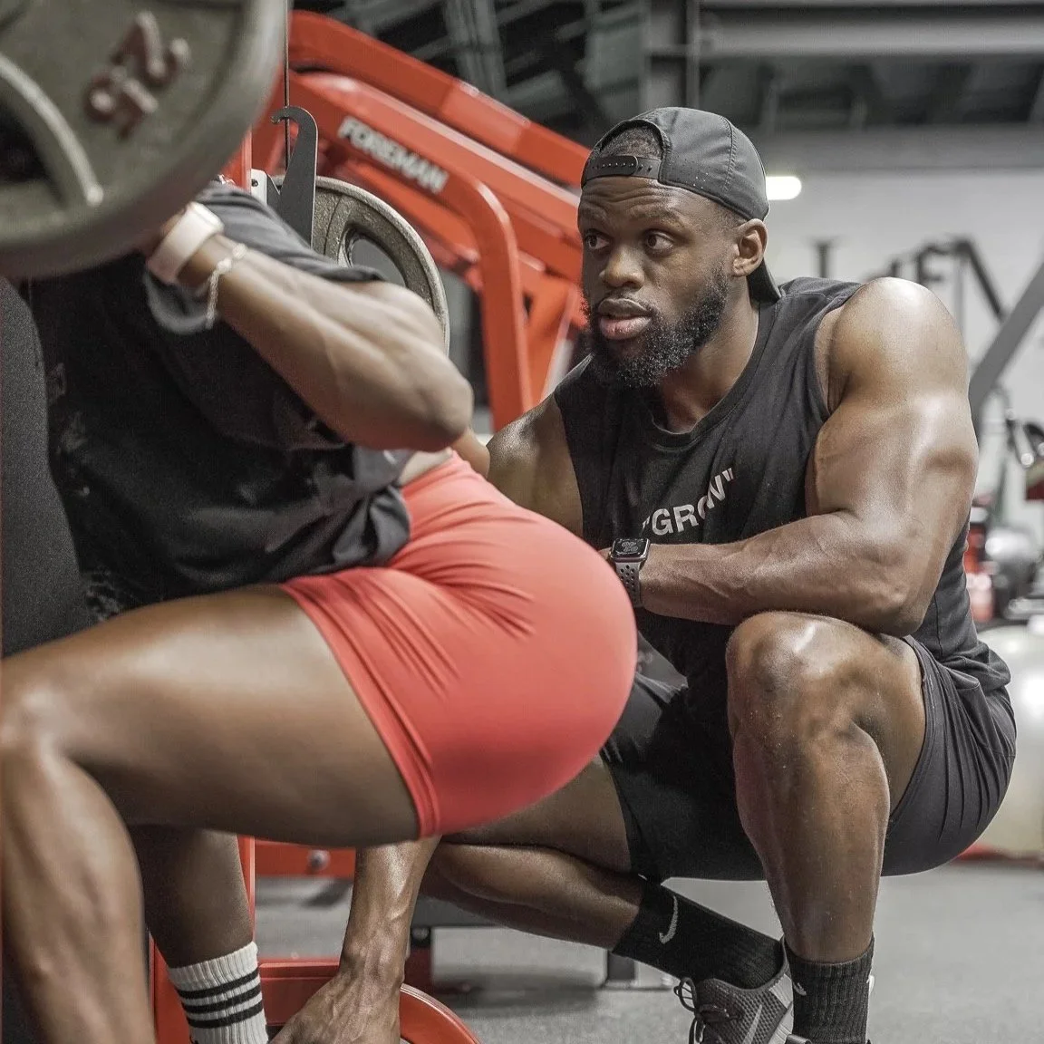 Coach Osamoje providing hands-on one-on-one training guidance during a leg workout at the gym, demonstrating proper squat form and strength coaching.