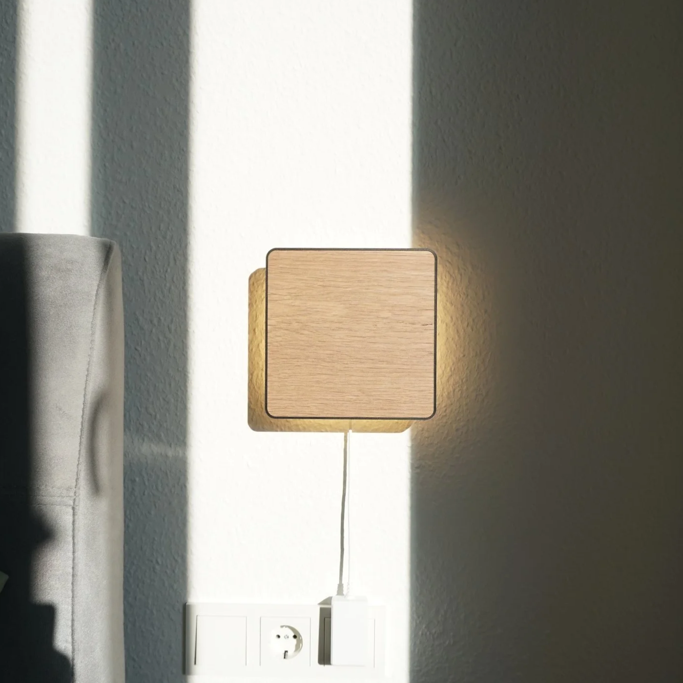 A wooden square-shaped lamp casting a soft glow on a white wall, with a power outlet and plug visible at the bottom.