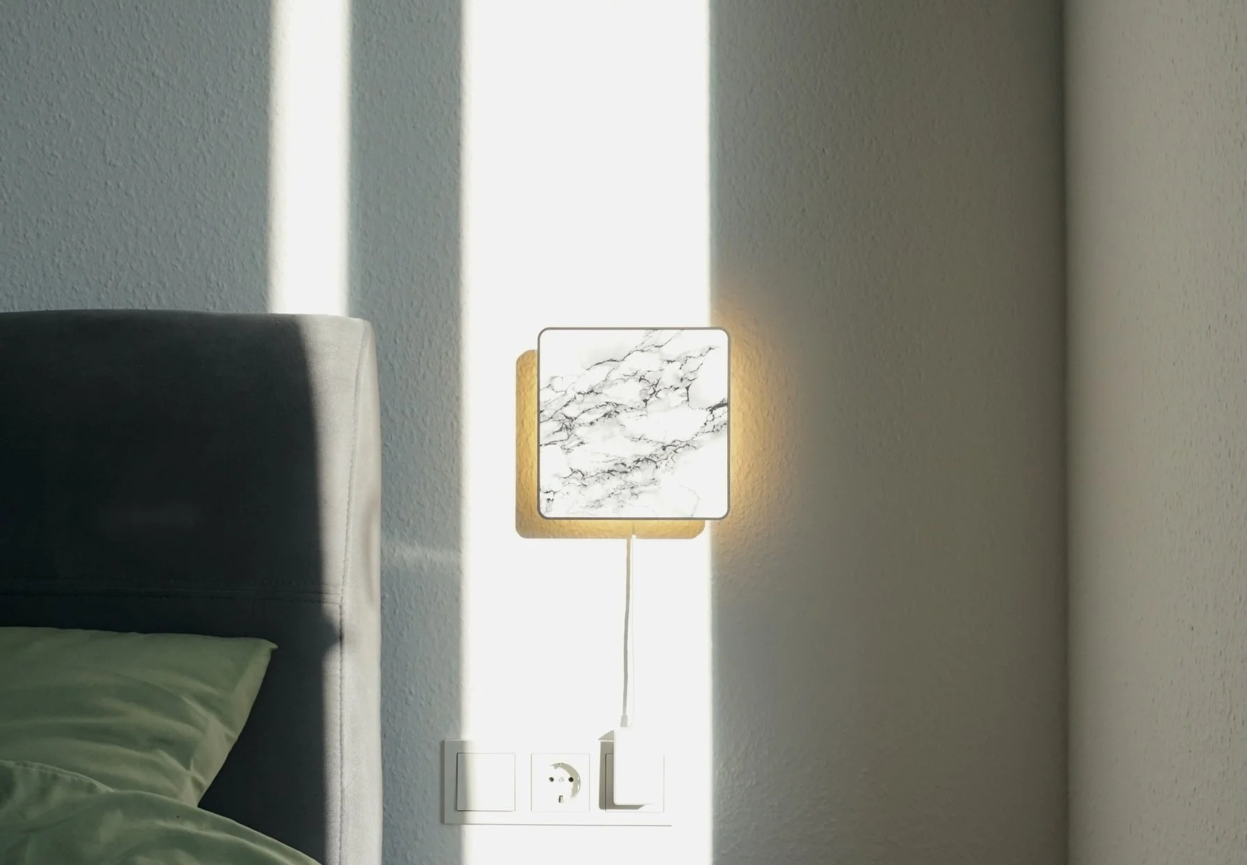 Wall-mounted square lamp with a marble pattern, glowing softly, next to a bed and electrical outlets.