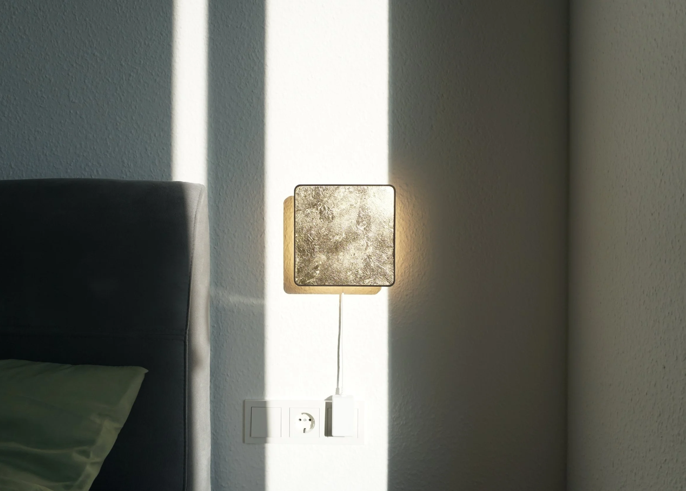A modern bedroom wall with a square textured wall light, an electrical outlet, and a bedside bed with a green pillow.
