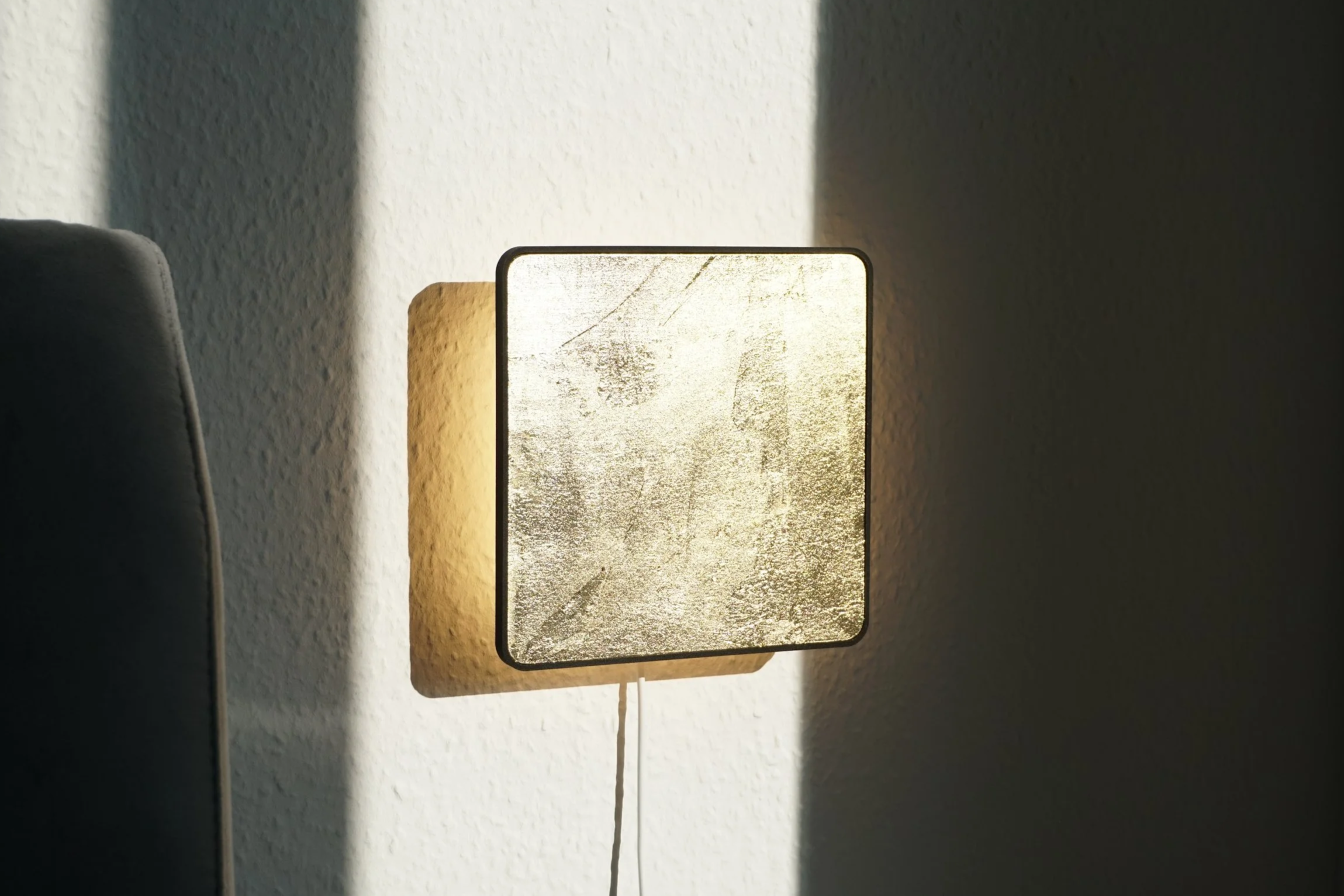 A modern wall-mounted lamp with a textured gold shade, casting a warm glow on a white wall, with a shadow on the right side of the image.