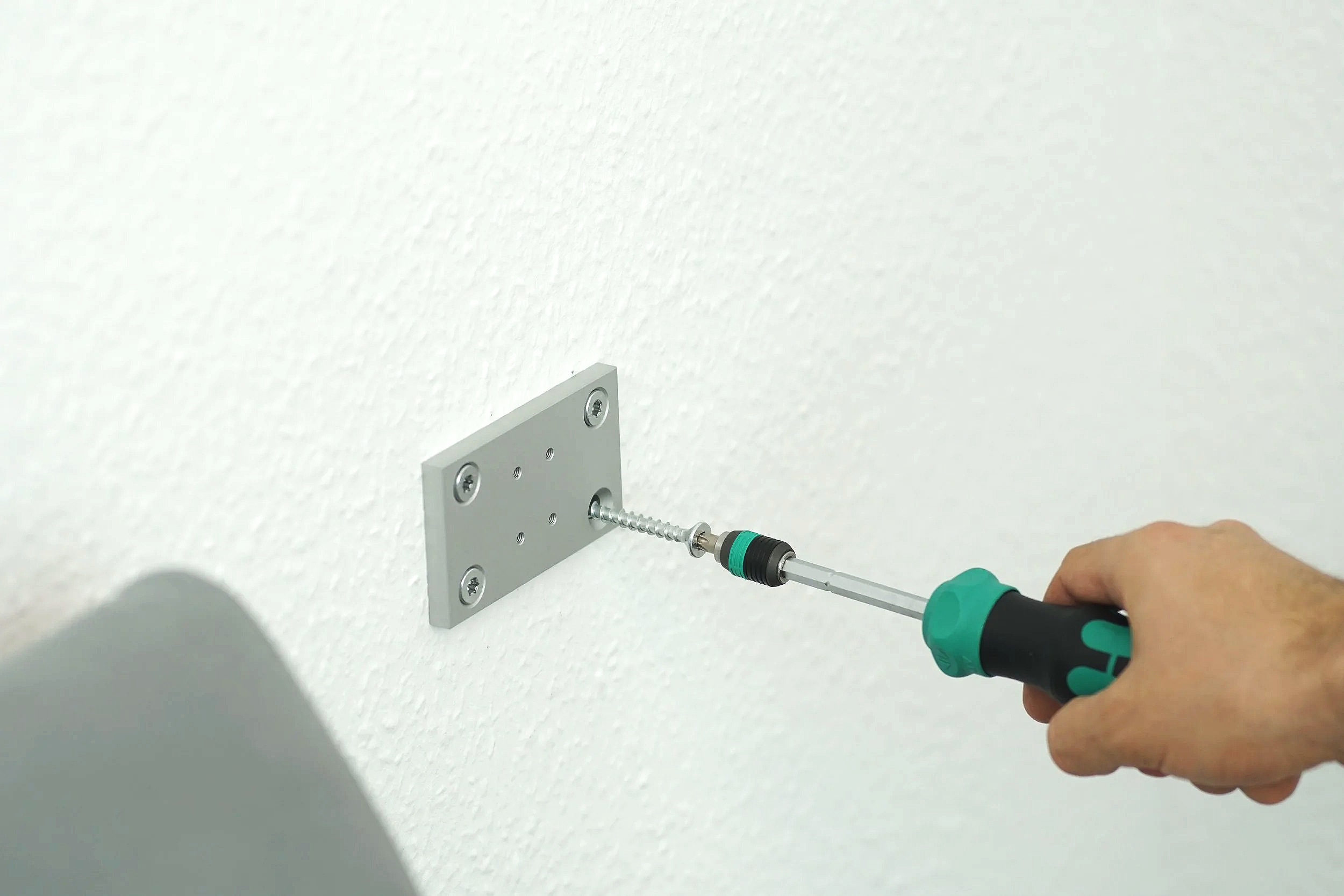 Person using a screwdriver to attach a metal plate with screws to a wall.