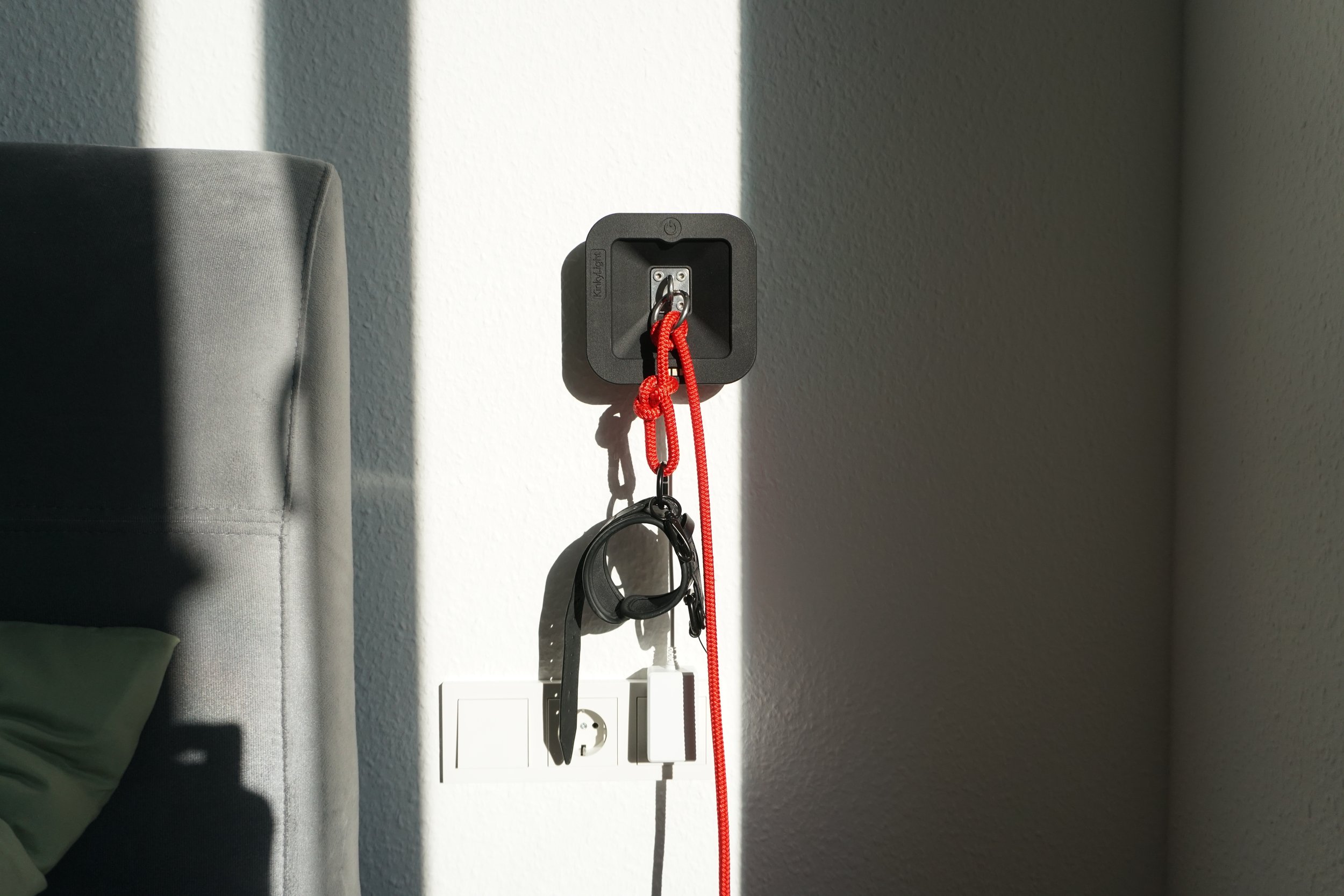 KinkyLight with kinky items hanging on it, including a red leash, black collar, and small black clip, near a gray upholstered headboard and a wall.