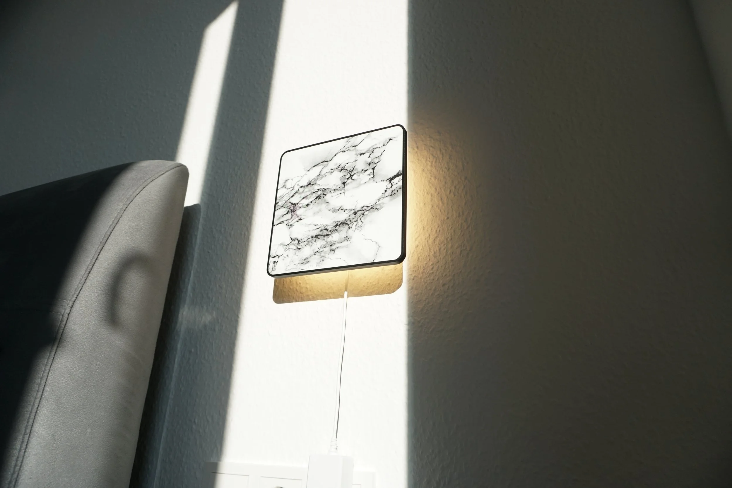 Wall-mounted bedside lamp with a marble pattern, illuminated and casting a shadow on a beige wall, positioned next to a gray upholstered headboard.