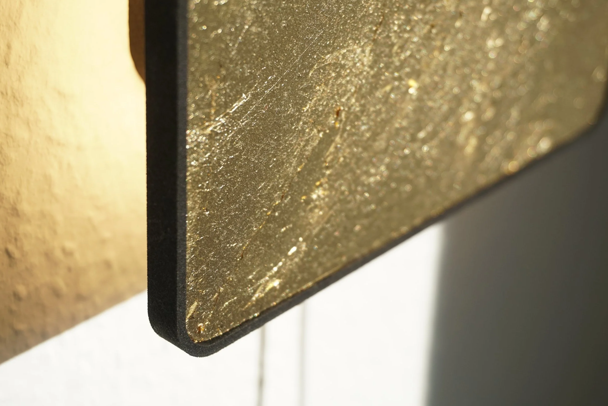 Close-up of a gold-colored surface with a rough, textured finish, and a black edge, illuminated by sunlight.
