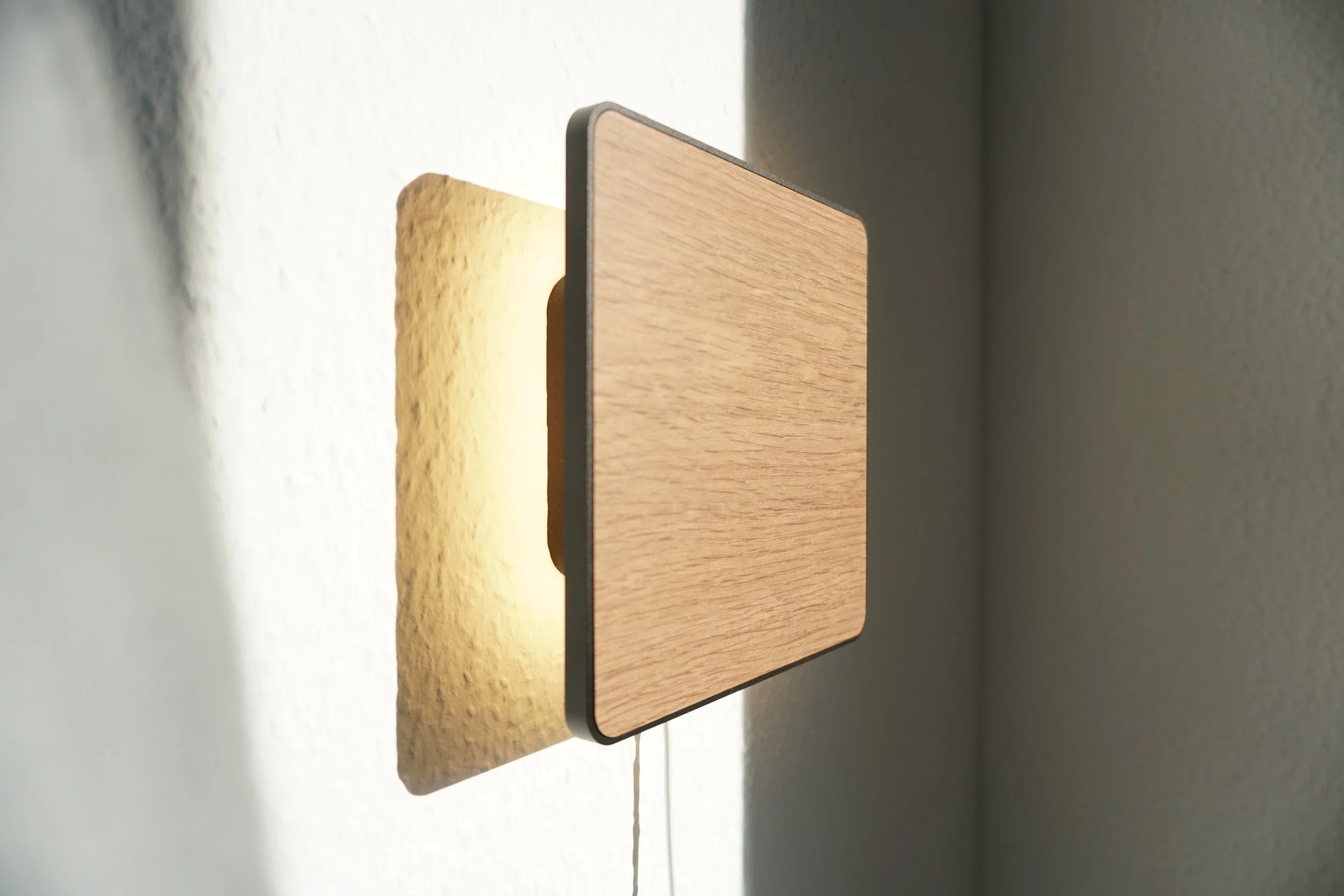 A wall-mounted bedside lamp with a minimalist design and a wooden textured front plate, casting a shadow on a textured white wall.