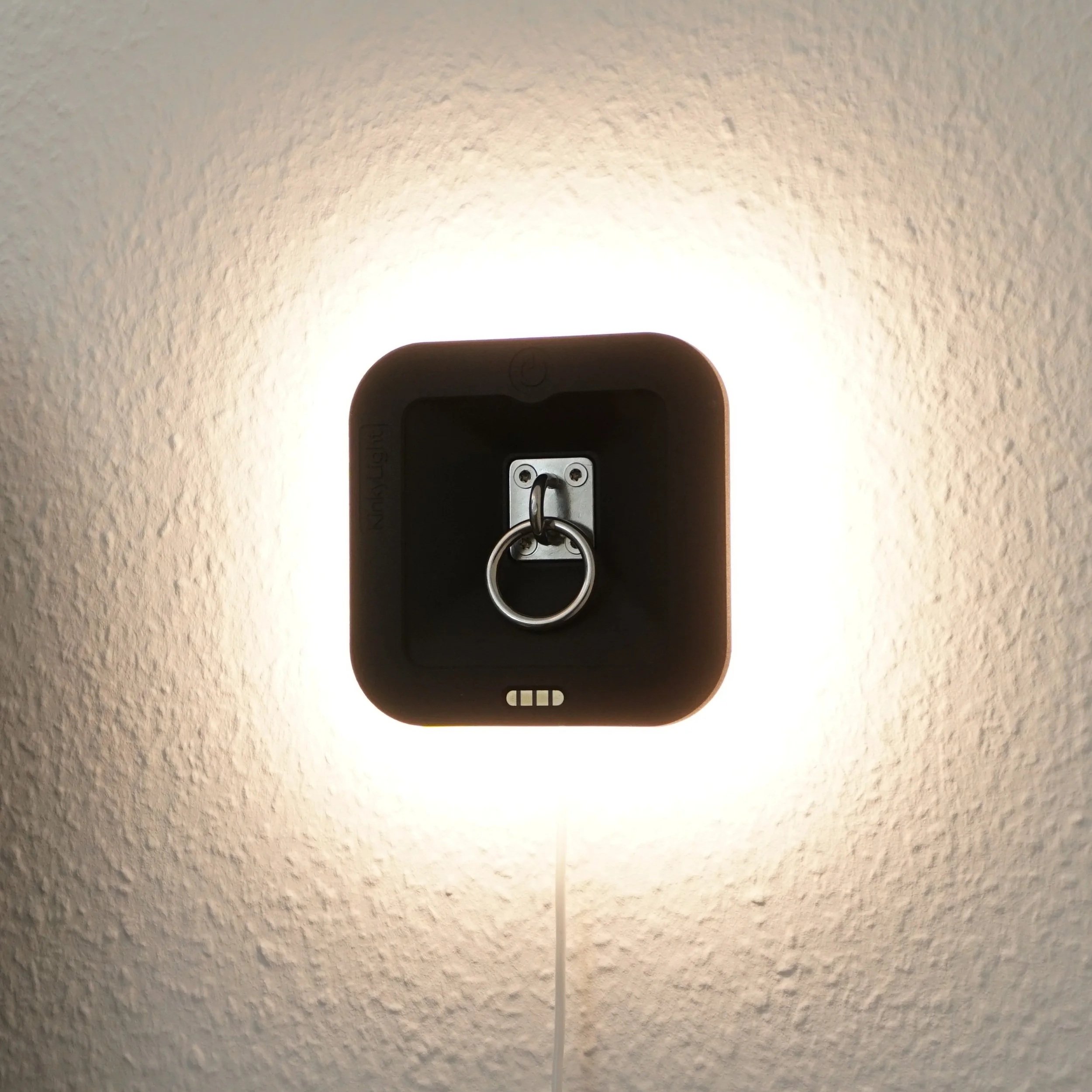 A wall-mounted night light with a ring tethered to a metal plate on an electrical outlet, casting a bright glow on a textured wall.