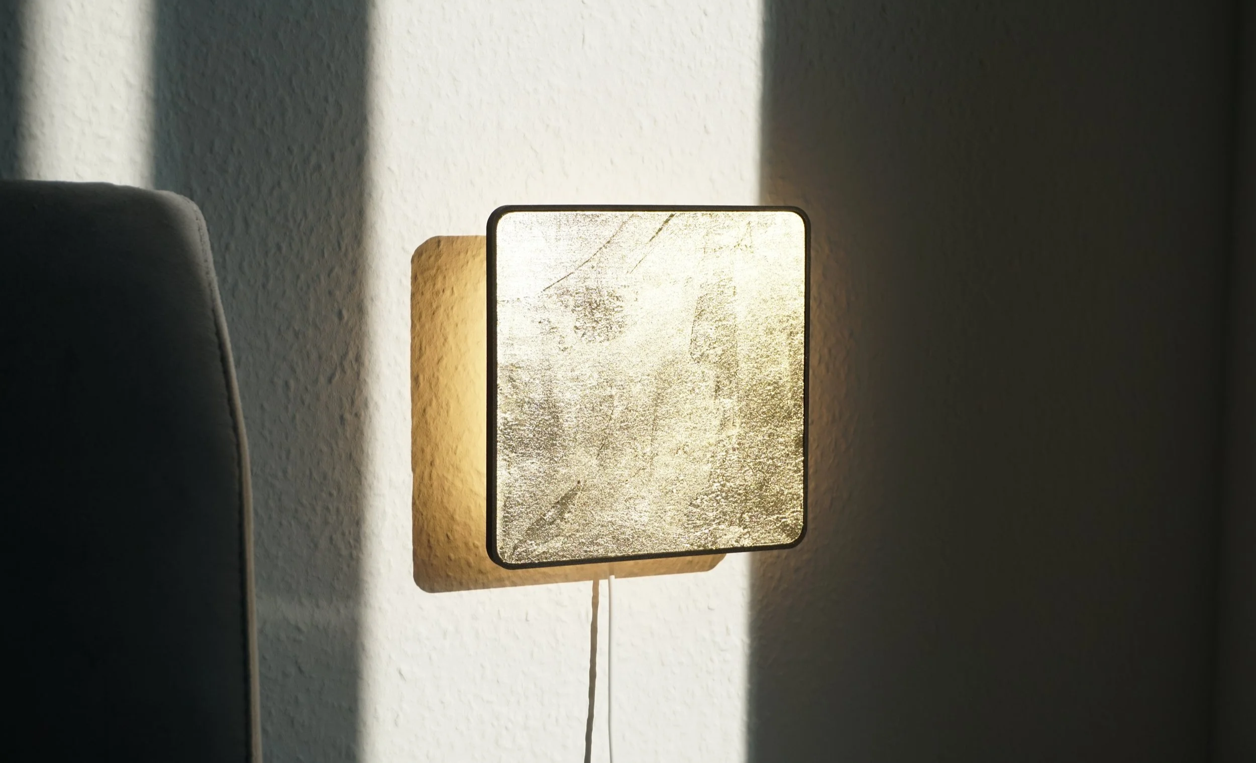 A square wall light with a textured glass surface casting a warm glow on a white wall, with part of a gray piece of furniture visible on the left.
