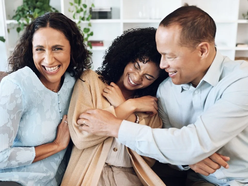 Three people sharing a joyful hug and laughter, two women and one man indoors with a bookshelf and plants in the background.