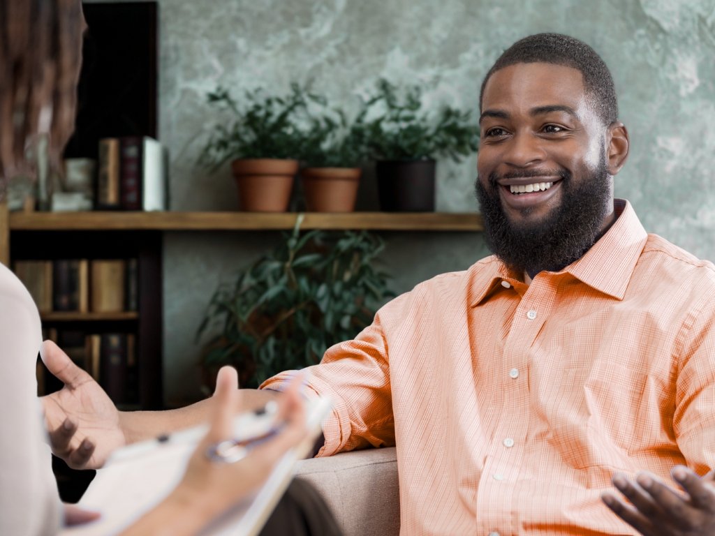 A man smiling and talking during a conversation, likely in a professional setting, with a woman holding a notepad in front of him.