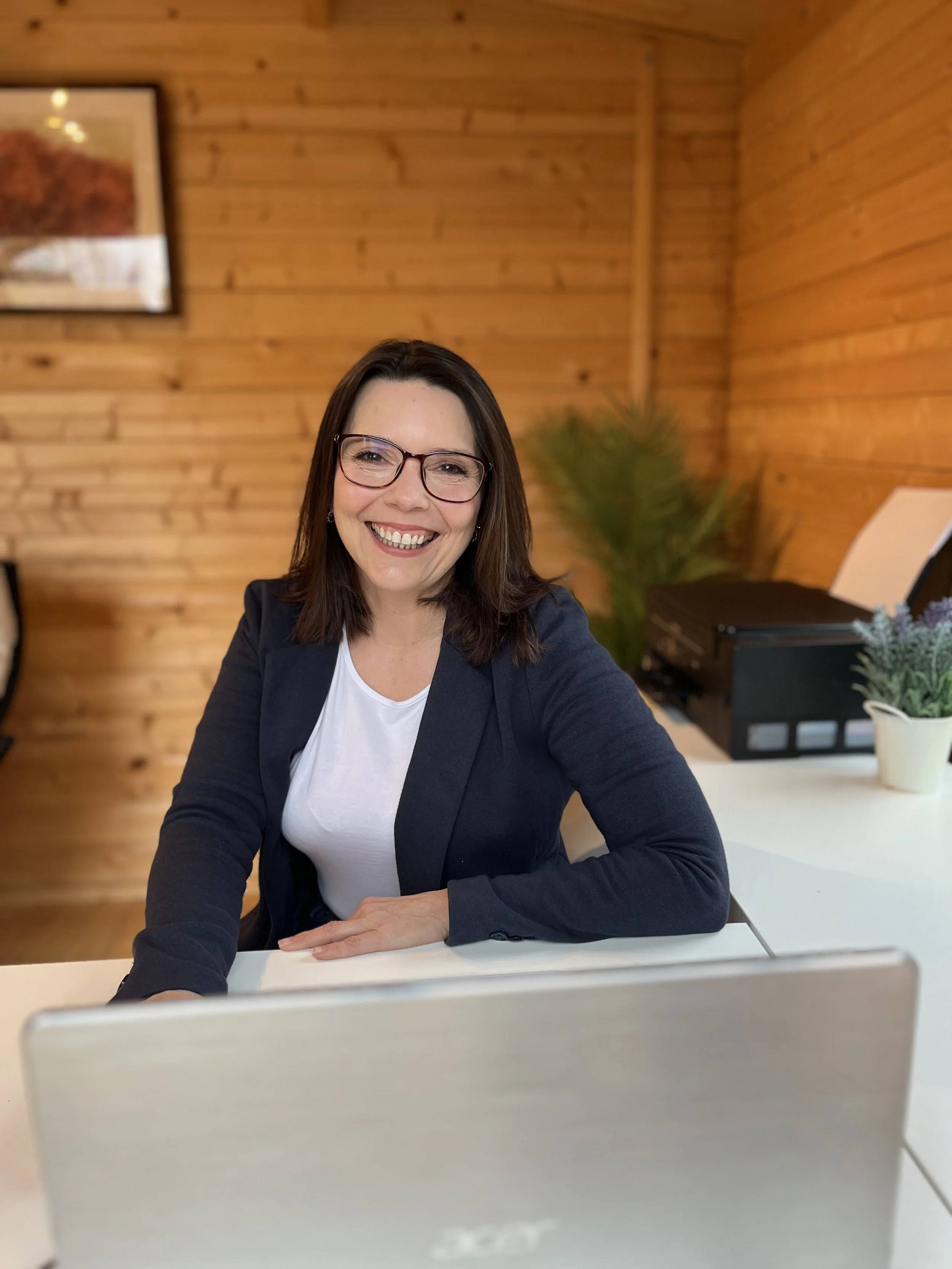 A woman with glasses smiling at a desk with a laptop in a wood-panelled office.