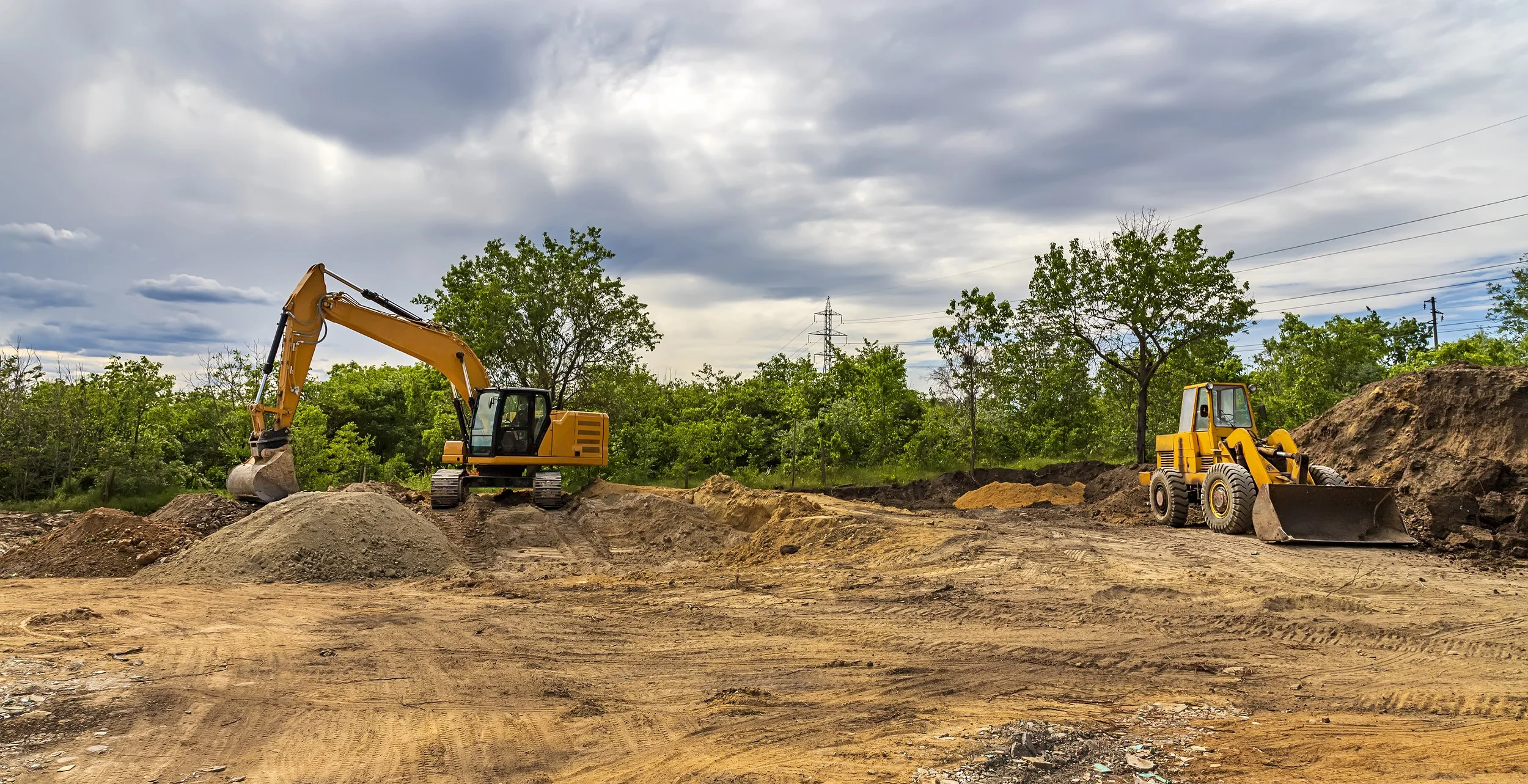 Construction site with two yellow excavators working on dirt and gravel, surrounded by green trees under a cloudy sky.