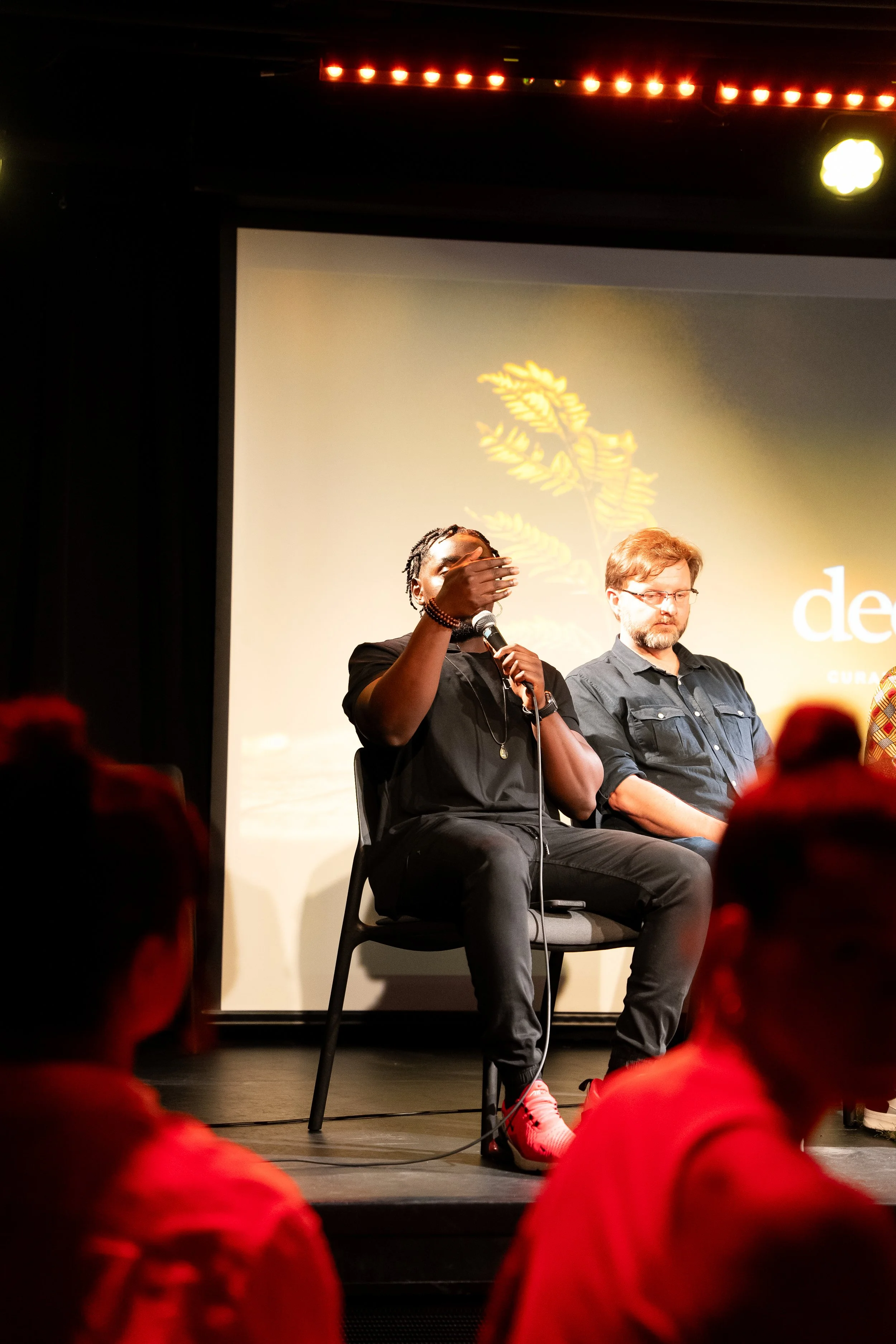Two men sitting on stage in front of a screen, with one speaking into a microphone and hand raised, audience members in front watching.