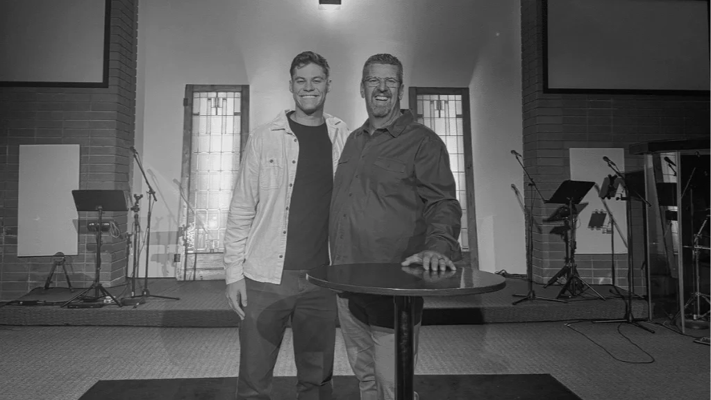 Two men standing together and smiling in a church or auditorium setting, with musical equipment and microphones in the background.