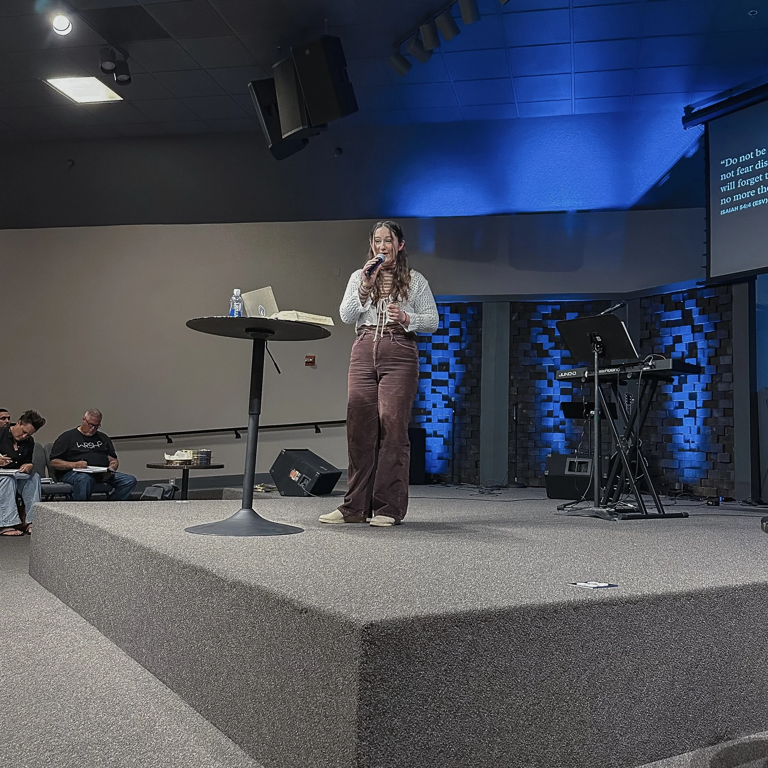 A woman stands on a stage, speaking into a microphone, with a music keyboard and a small table with a water bottle and open notebook beside her. There are audience members seated in the background.