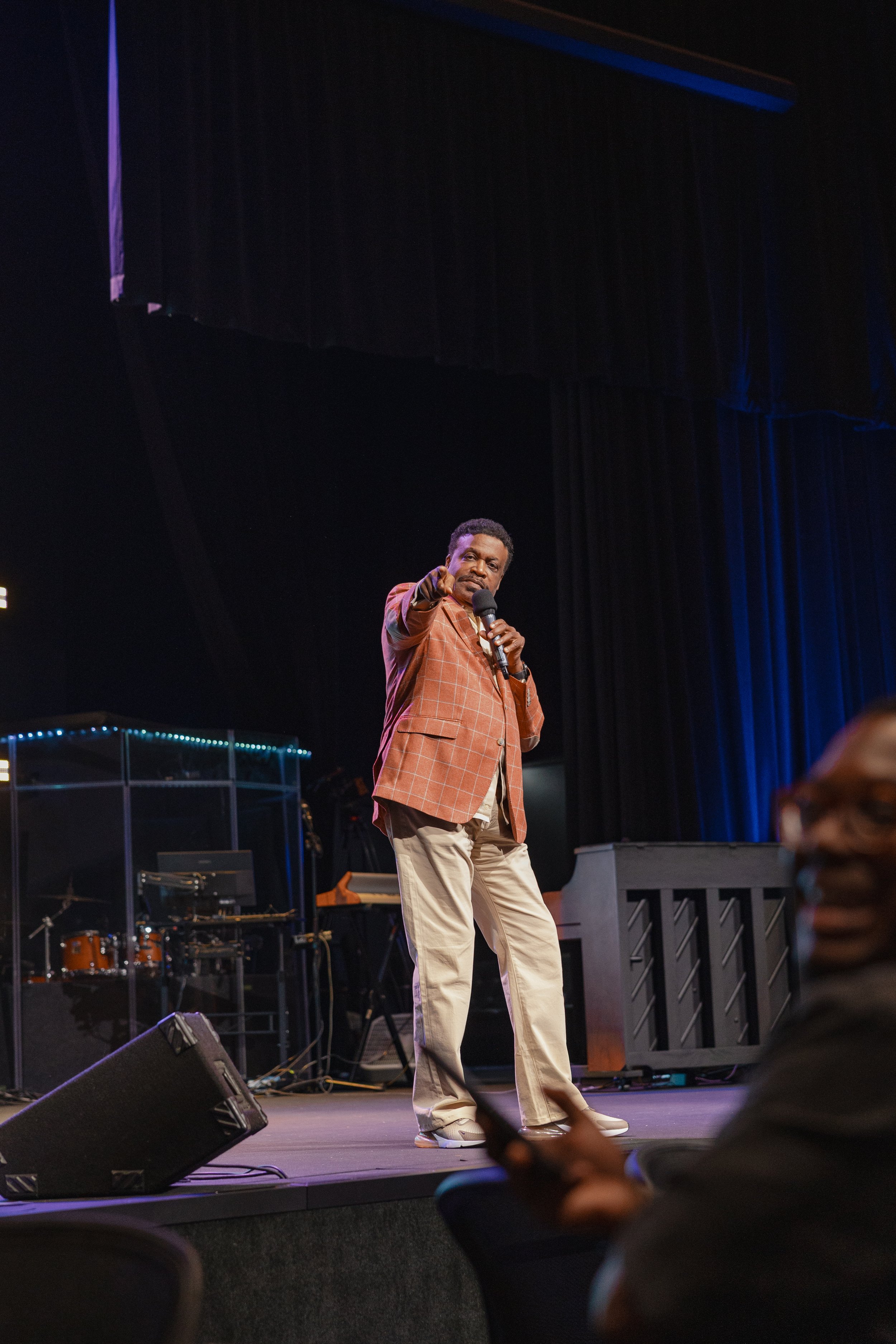A man in a brown checkered blazer and beige pants is on stage, holding a microphone and pointing towards the audience during a performance or speech, with musical instruments in the background.