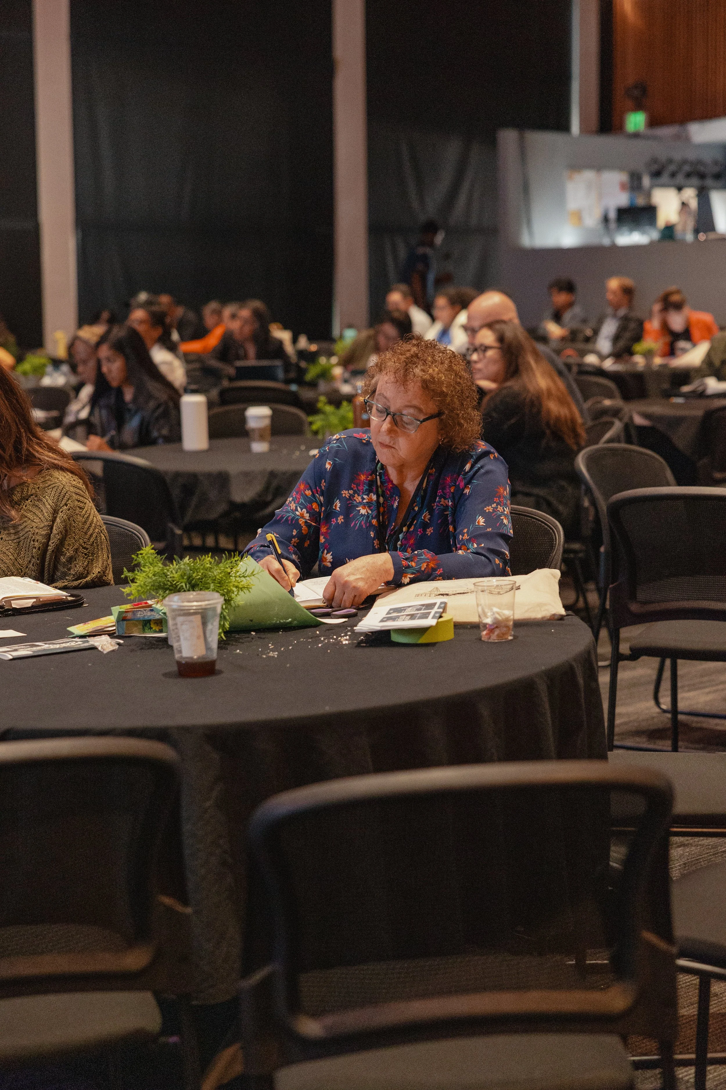 A woman with curly hair, glasses, and a floral blouse sitting at a round table during a conference or seminar, looking at documents and writing.