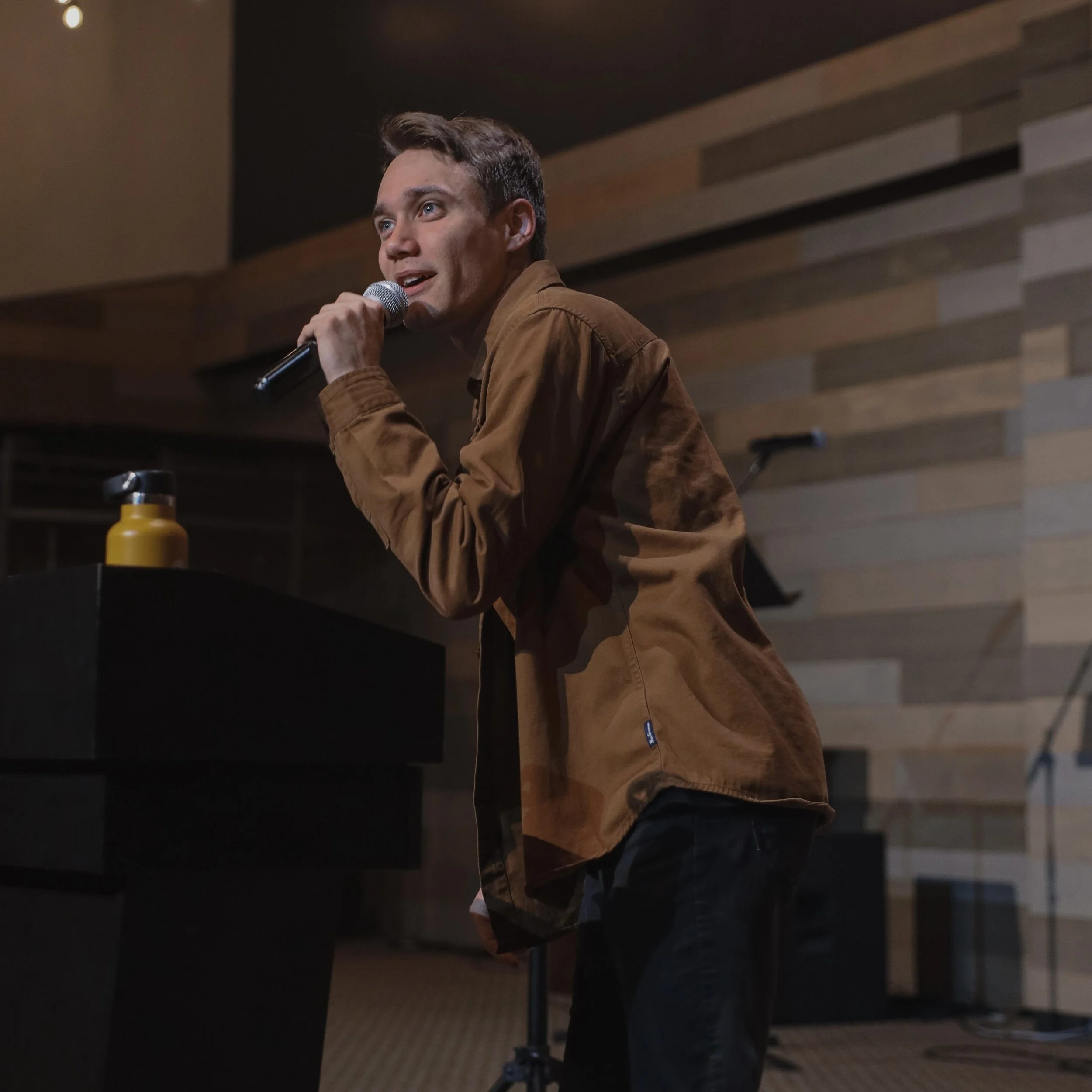 Man stand with a microphone on stage, wearing a brown jacket and black pants, with a yellow water bottle on a black podium nearby.