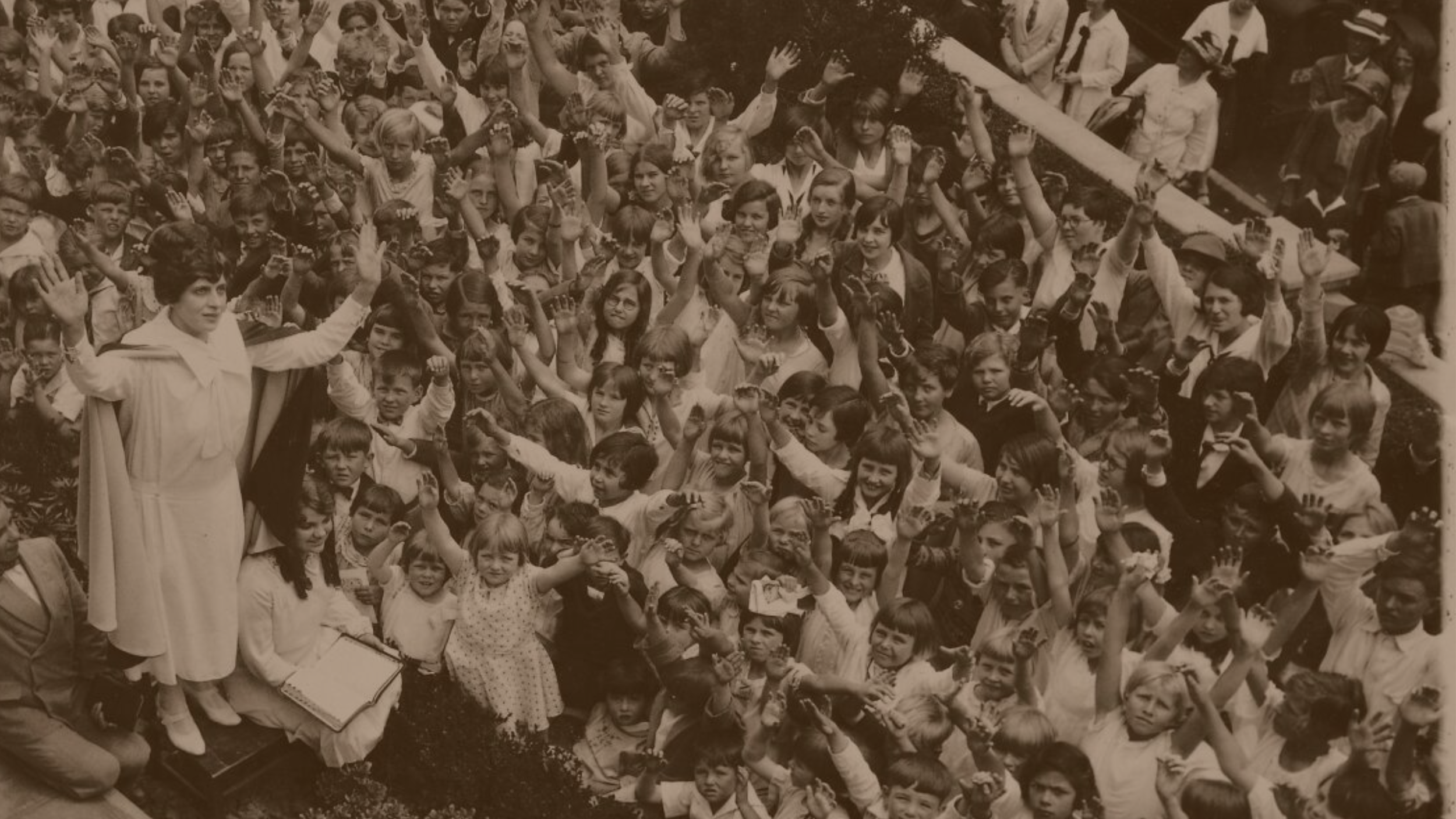 Black and white historical photo of a large crowd of children and women gathered outdoors, some raising their hands, with a woman in a white dress and cape standing on a platform in front of them.