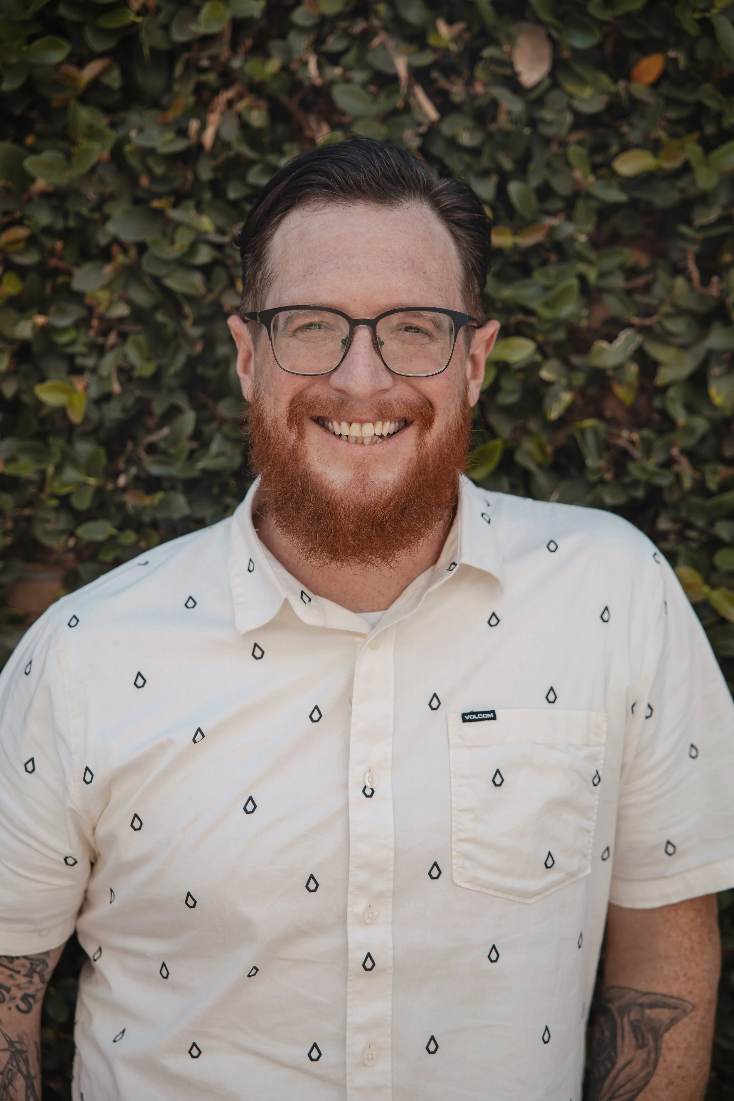 A smiling man with glasses and a beard, wearing a white short-sleeved button-up shirt with small black diamond patterns, standing in front of a leafy background.