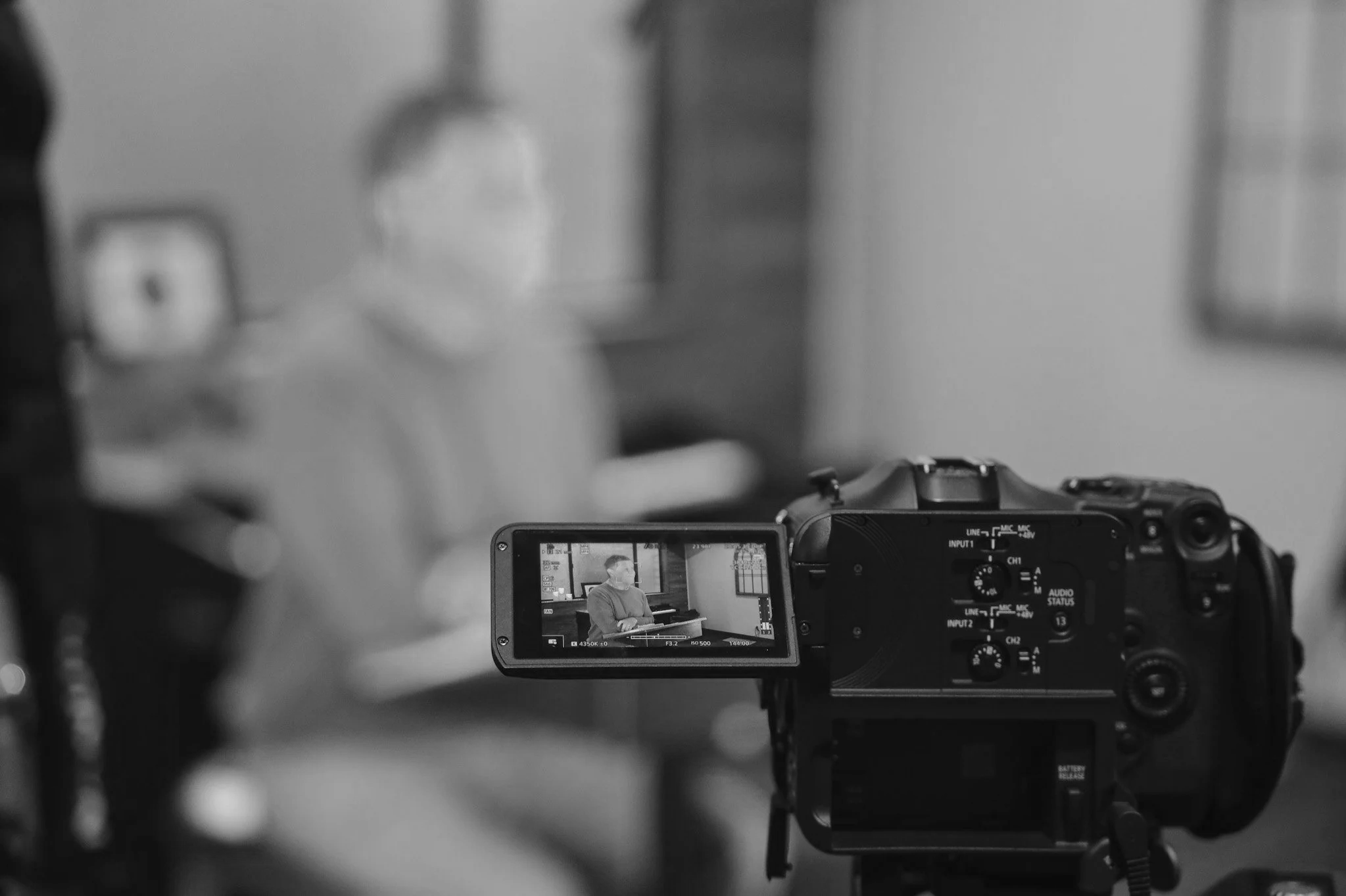 A person being filmed with a professional video camera in a room, with the camera's LCD screen showing a man sitting at a desk.