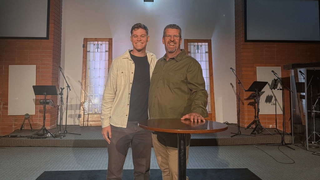 Two smiling men standing on a stage with musical equipment and microphones, one with his hand on a round table.