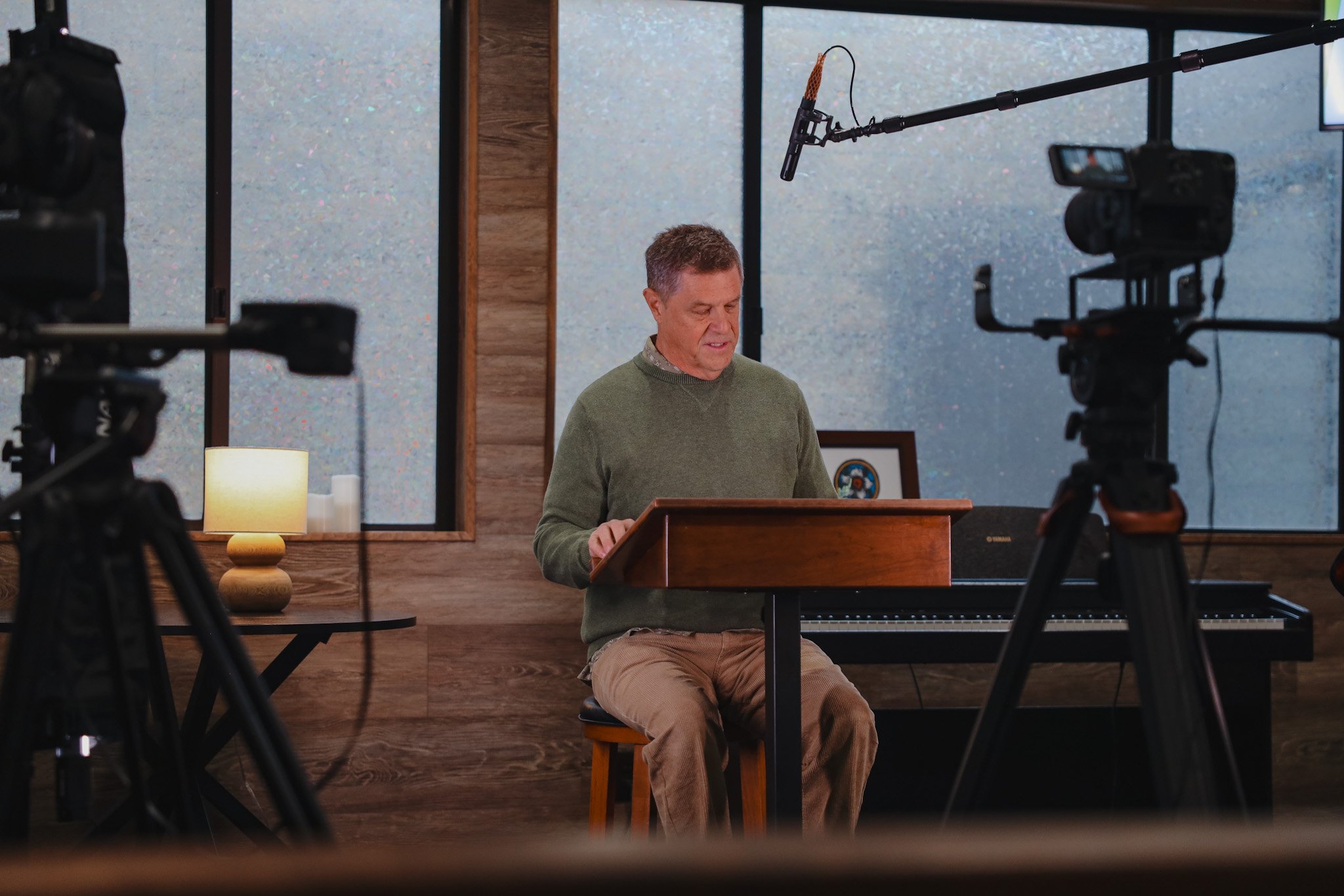 A man in a green sweater sitting at a wooden table with a microphone overhead, filming equipment in the foreground, behind him a piano, a small table with a lamp, and a window with frosted glass.