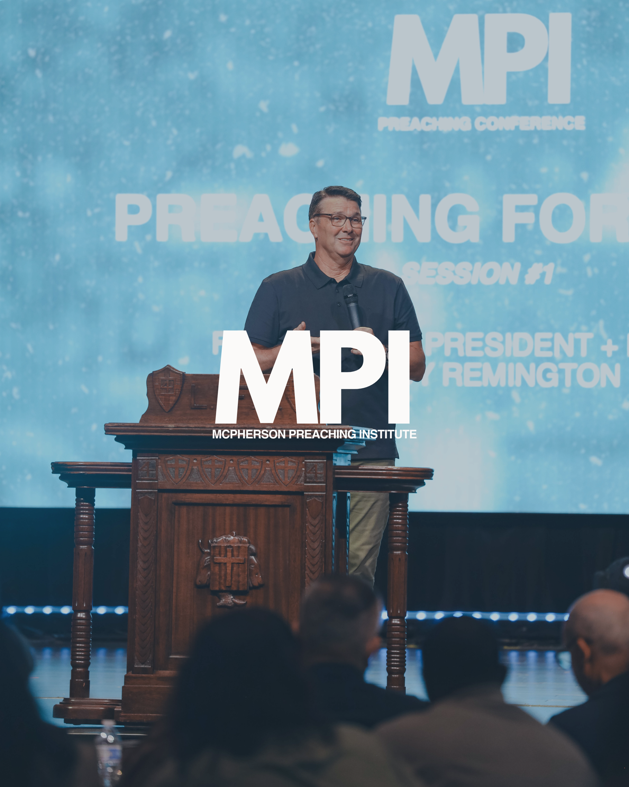 A man standing at a wooden podium speaking into a microphone during a conference, with a blue backdrop displaying 'MPI Preaching Conference' and 'Session #1'. Audience members are visible in the foreground.