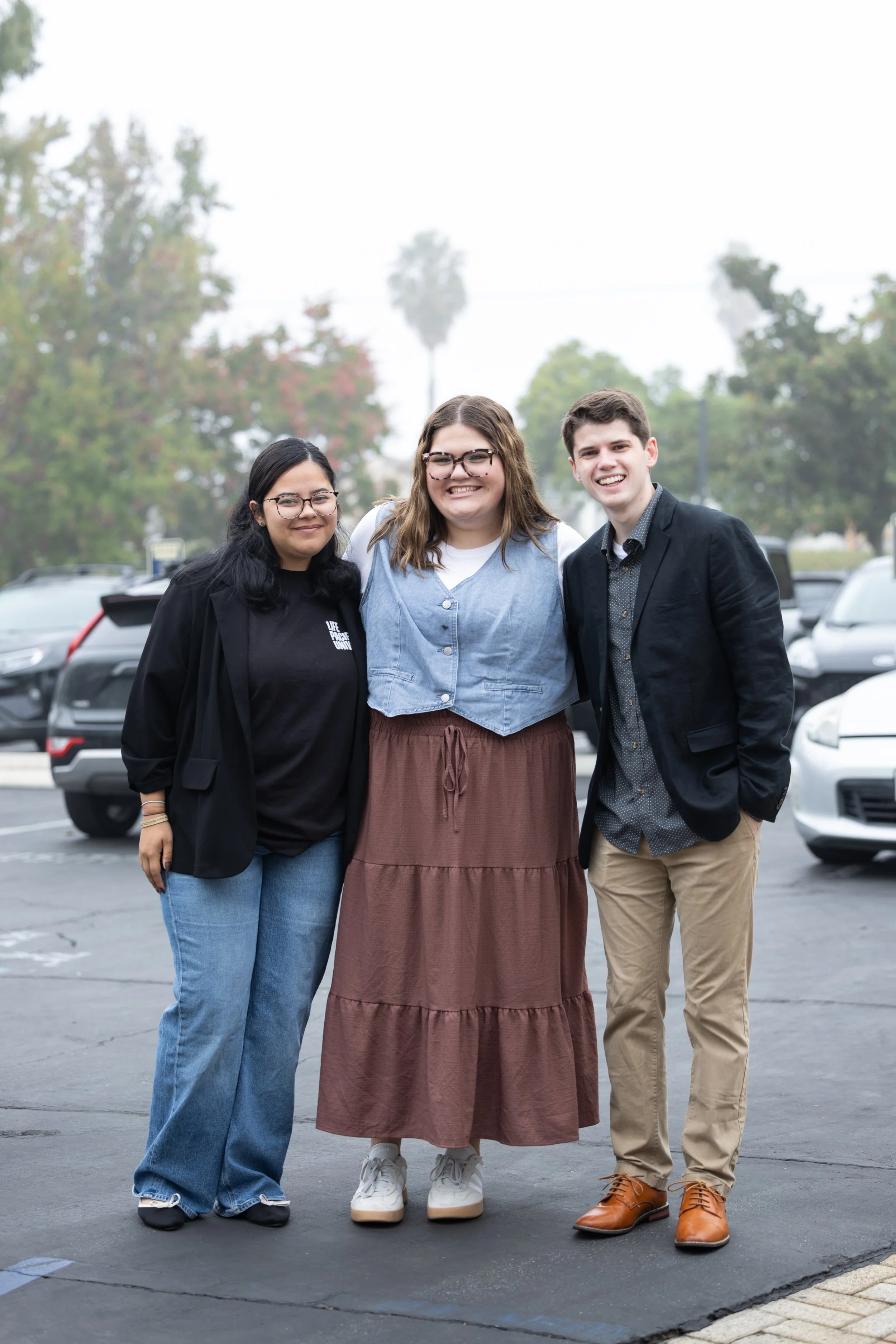 Three young adults standing together outdoors in a parking lot, smiling at the camera, with trees and cars in the background.
