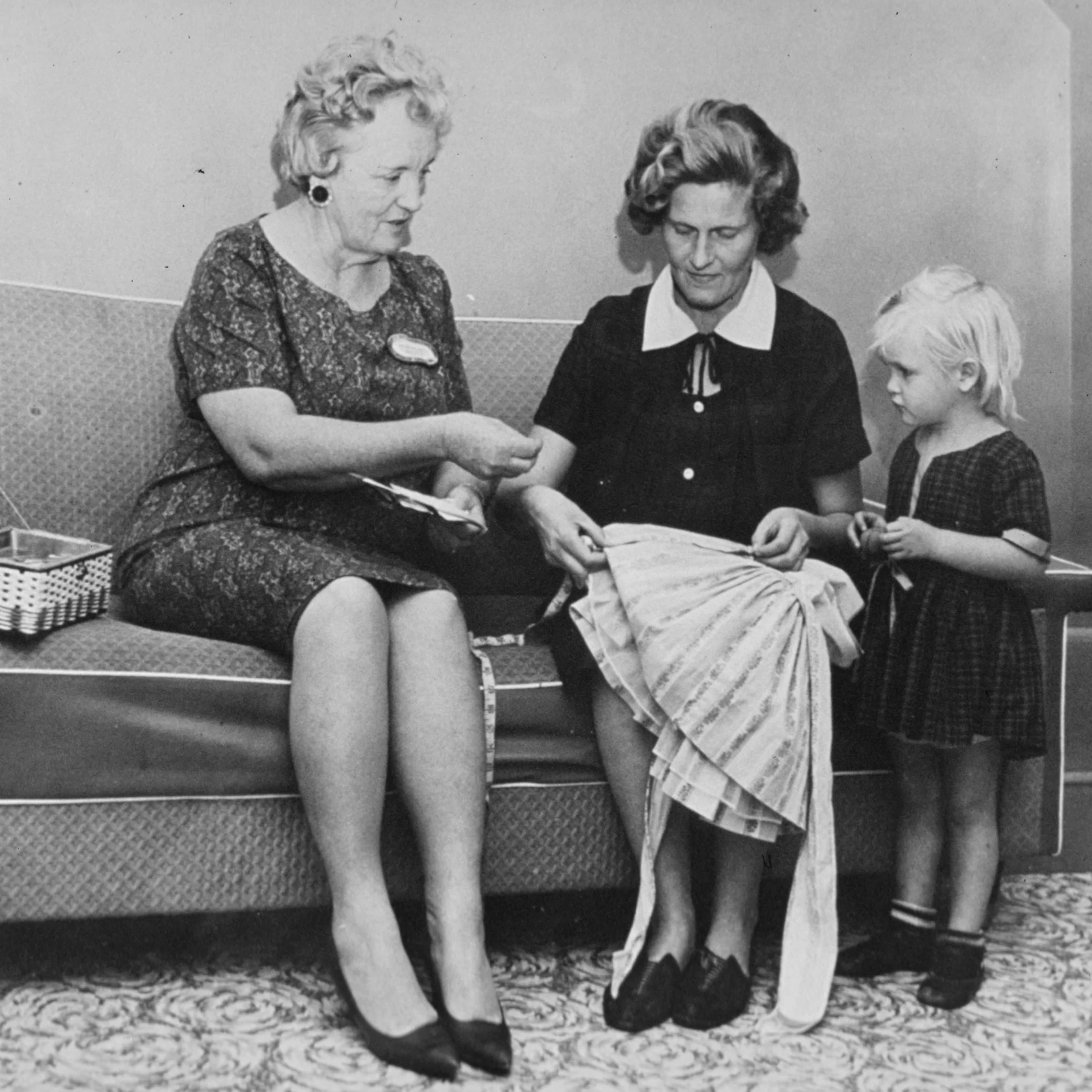 Trois femmes et une jeune fille dans une pièce, avec deux femmes assises et une enfant se tenant debout, en train de regarder une couture ou un atelier de couture.