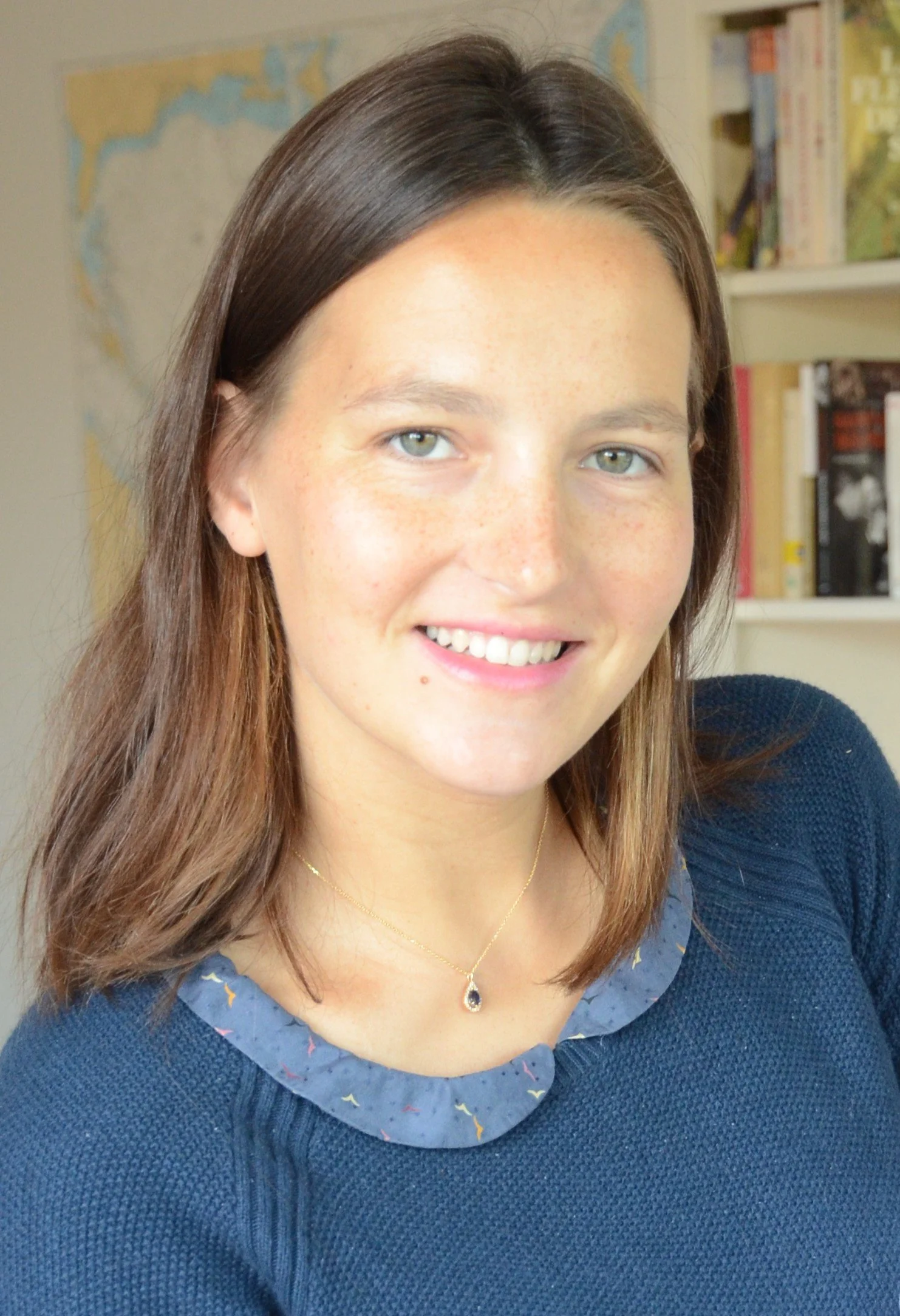 Jeune femme avec des cheveux bruns, souriante, portant un collier avec pendentif, en arrière-plan une étagère avec des livres et une carte du monde.