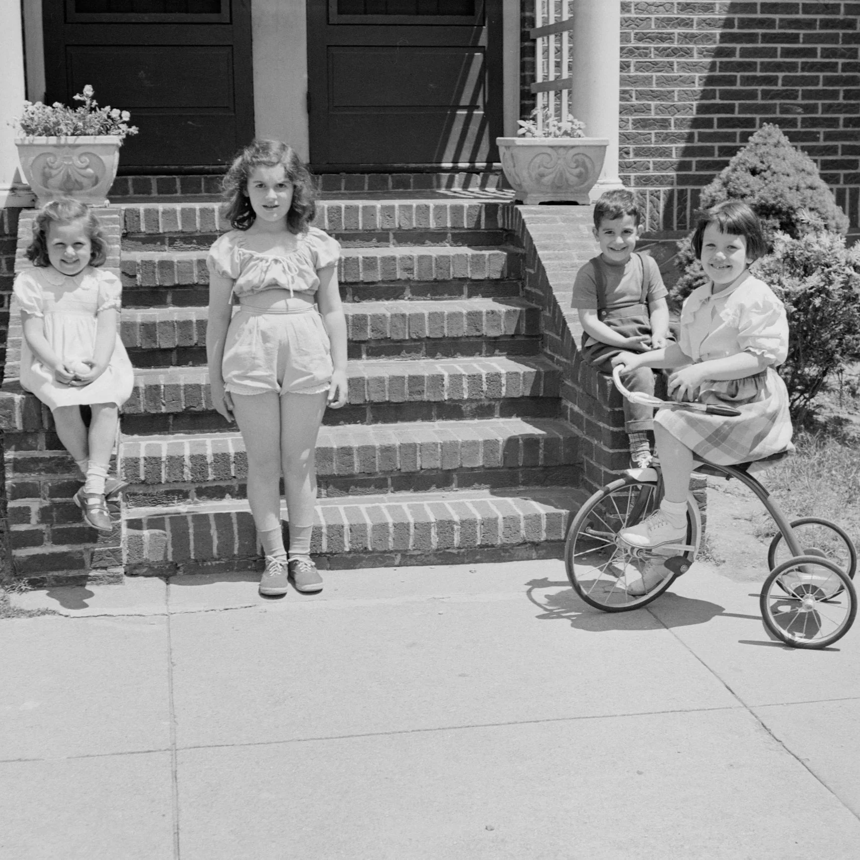 Quatre enfants posant devant une maison en briques, deux de chaque côté d'escalier. Deux filles en robes courtes et deux garçons, dont une fille sur un tricycle.