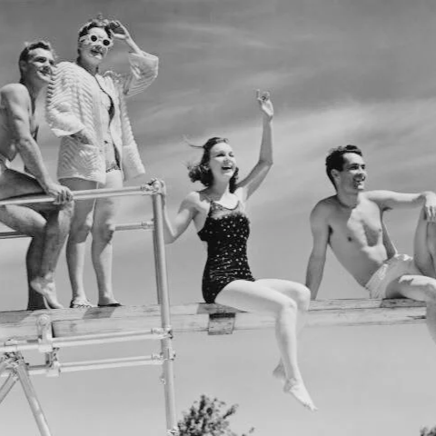 Quatre personnes profitant d'une journée à la plage, assises sur un quai ou une plateforme en bois, avec un ciel nuageux en fond, en noir et blanc.