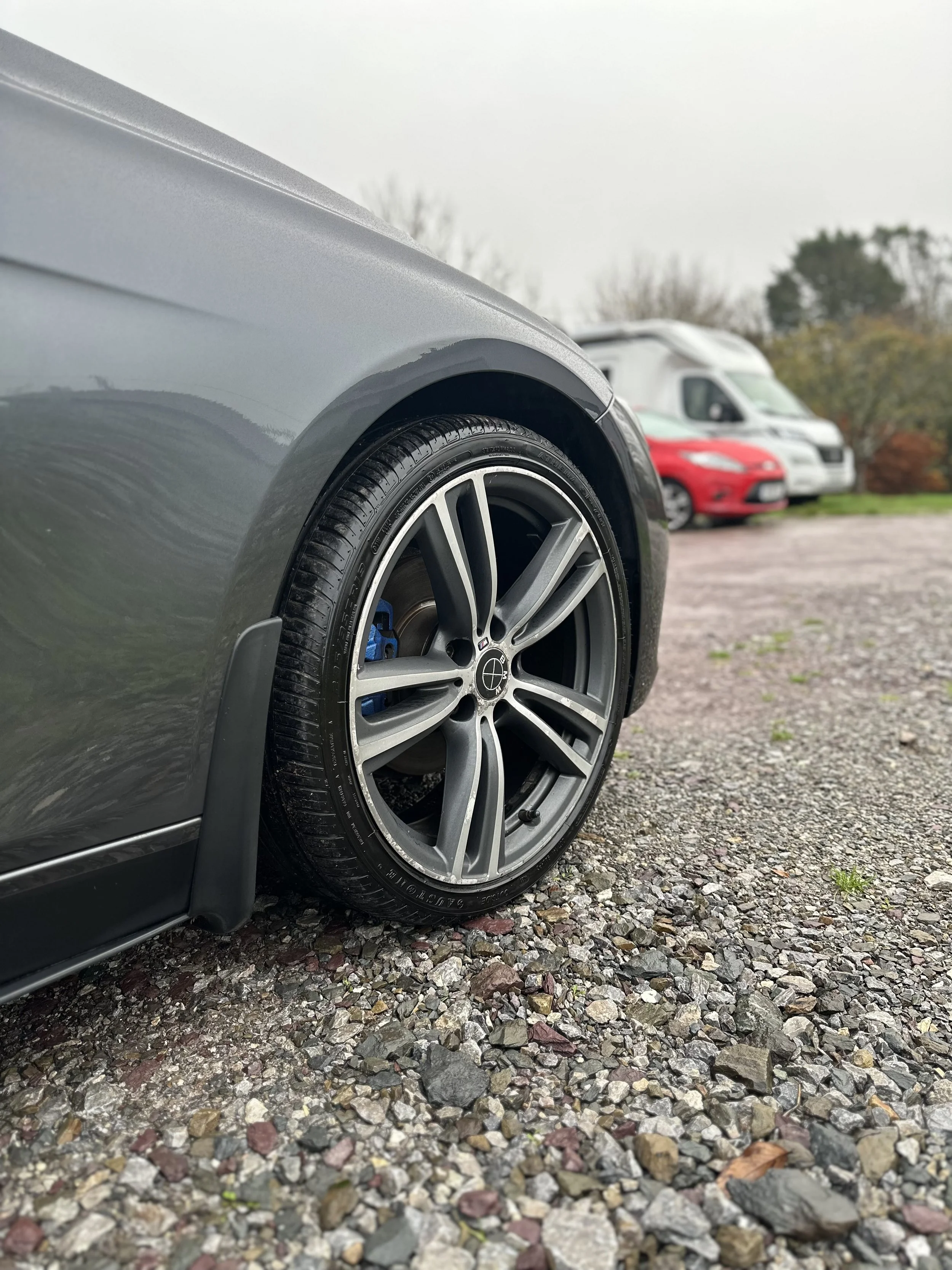 Close-up of a gray car's front wheel with a low profile tire and alloy rim, parked on a gravel surface, with two vehicles and some trees in the background.