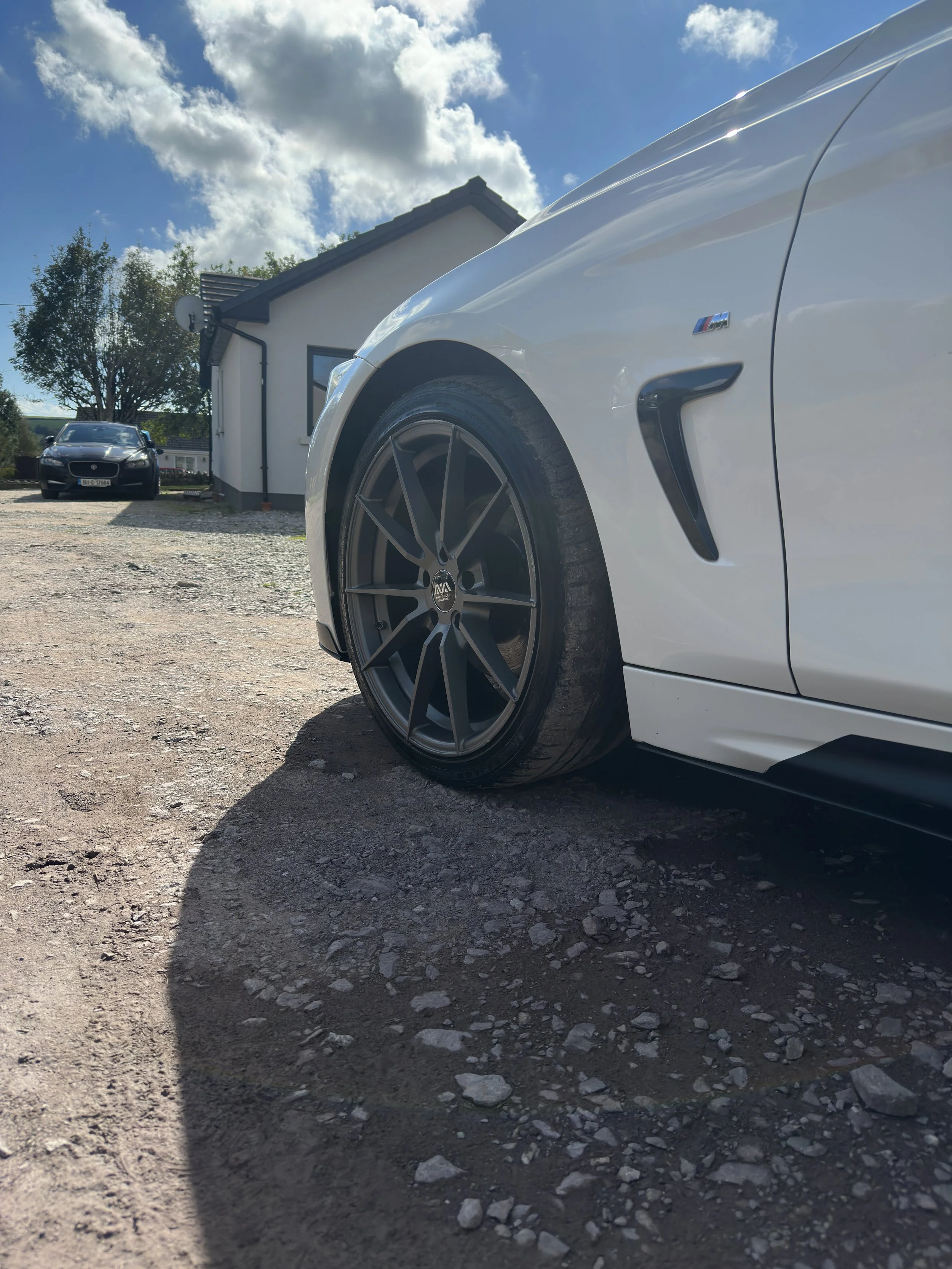 Close-up of a white sporty car's front tire and side vent, with a house and another car visible in the background under a partly cloudy sky.