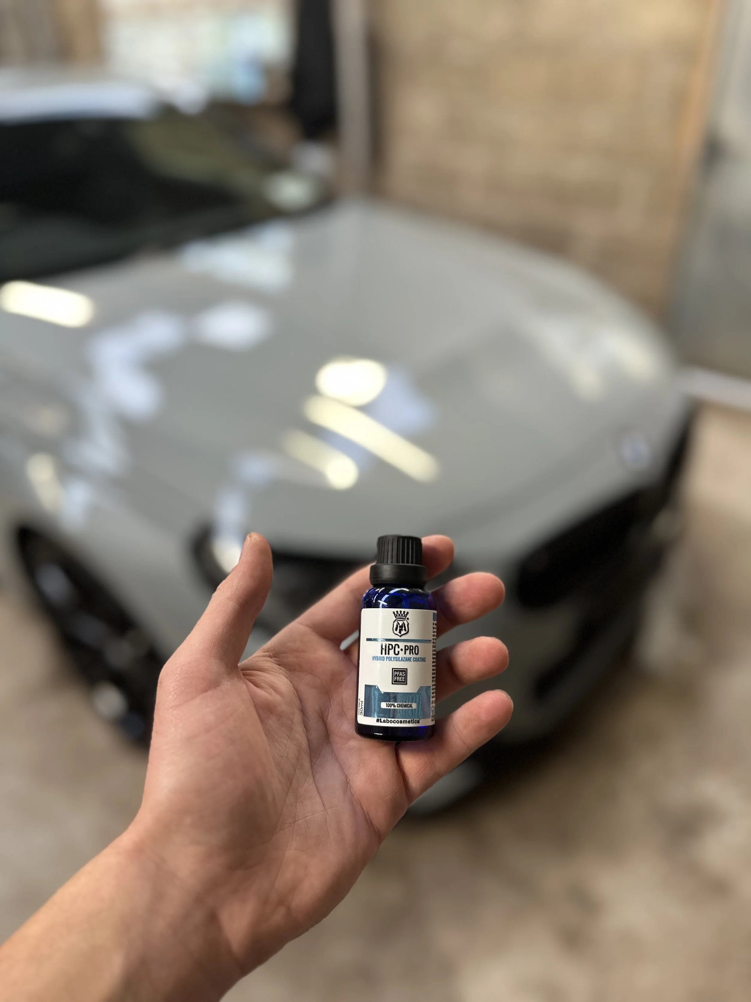 Person holding a small blue bottle labeled HPC-PRO in front of a blurred white car in a garage.