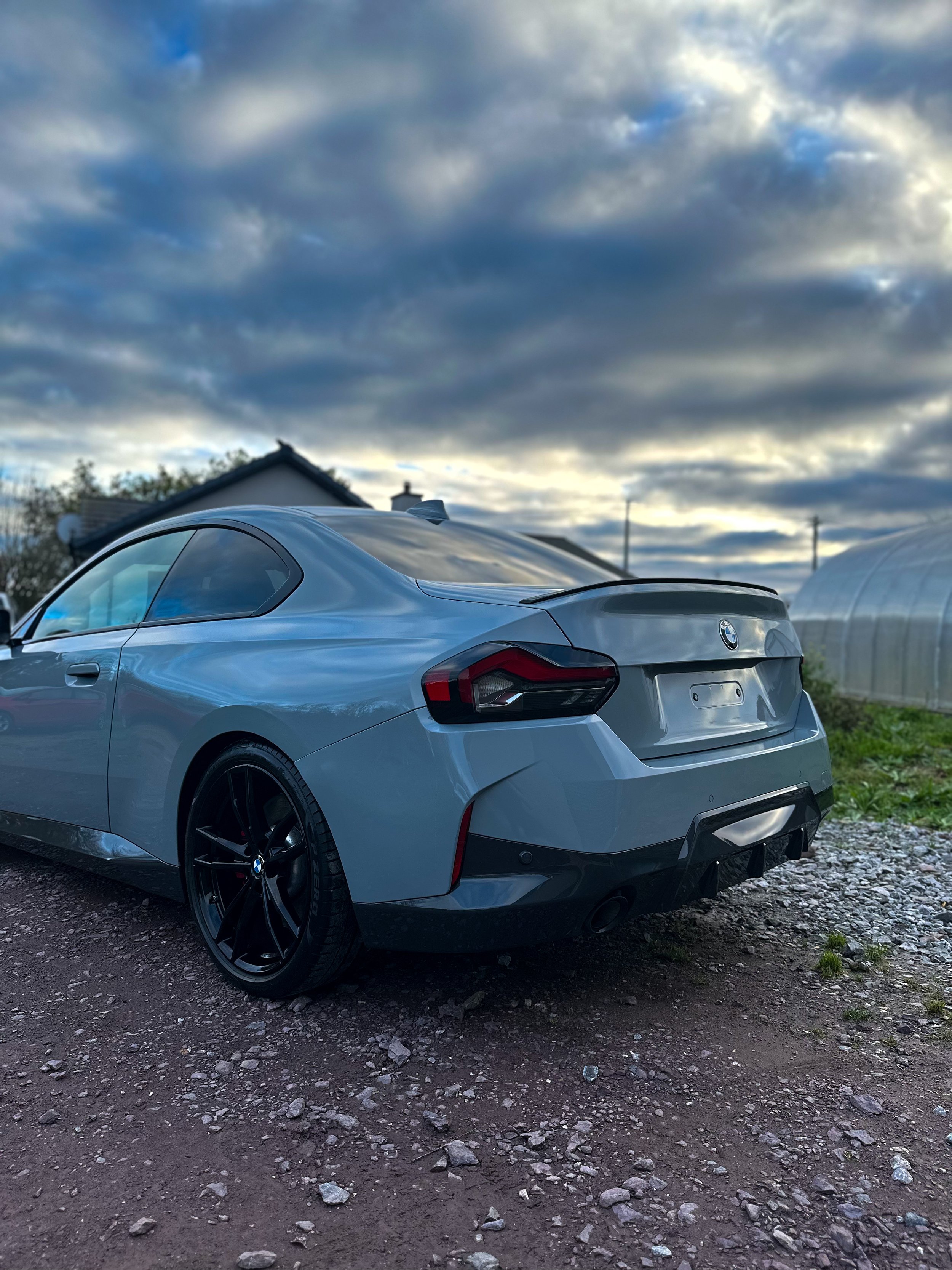 A silver BMW coupe parked on a gravel surface outdoors with a cloudy sky in the background.