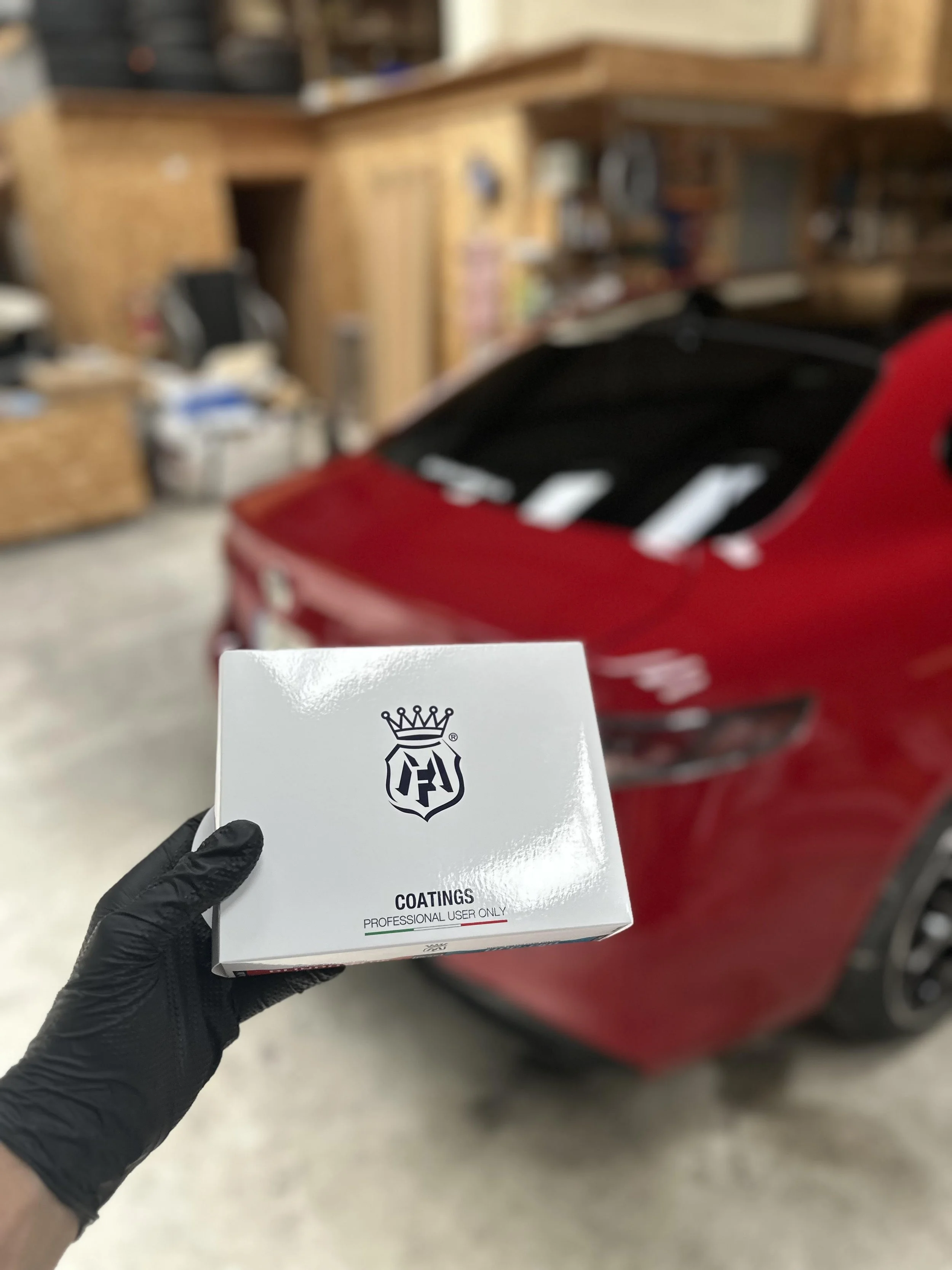 Hand wearing a black glove holding a box labeled 'Coatings' in front of a red car inside a garage.