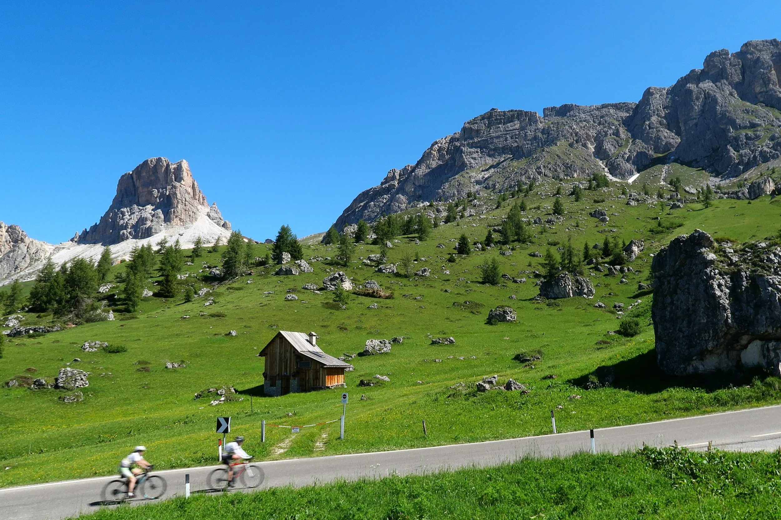 Scenic mountain landscape with rocky peaks, green grassy slopes, small trees, a wooden hut, and people riding bicycles on a road in the foreground