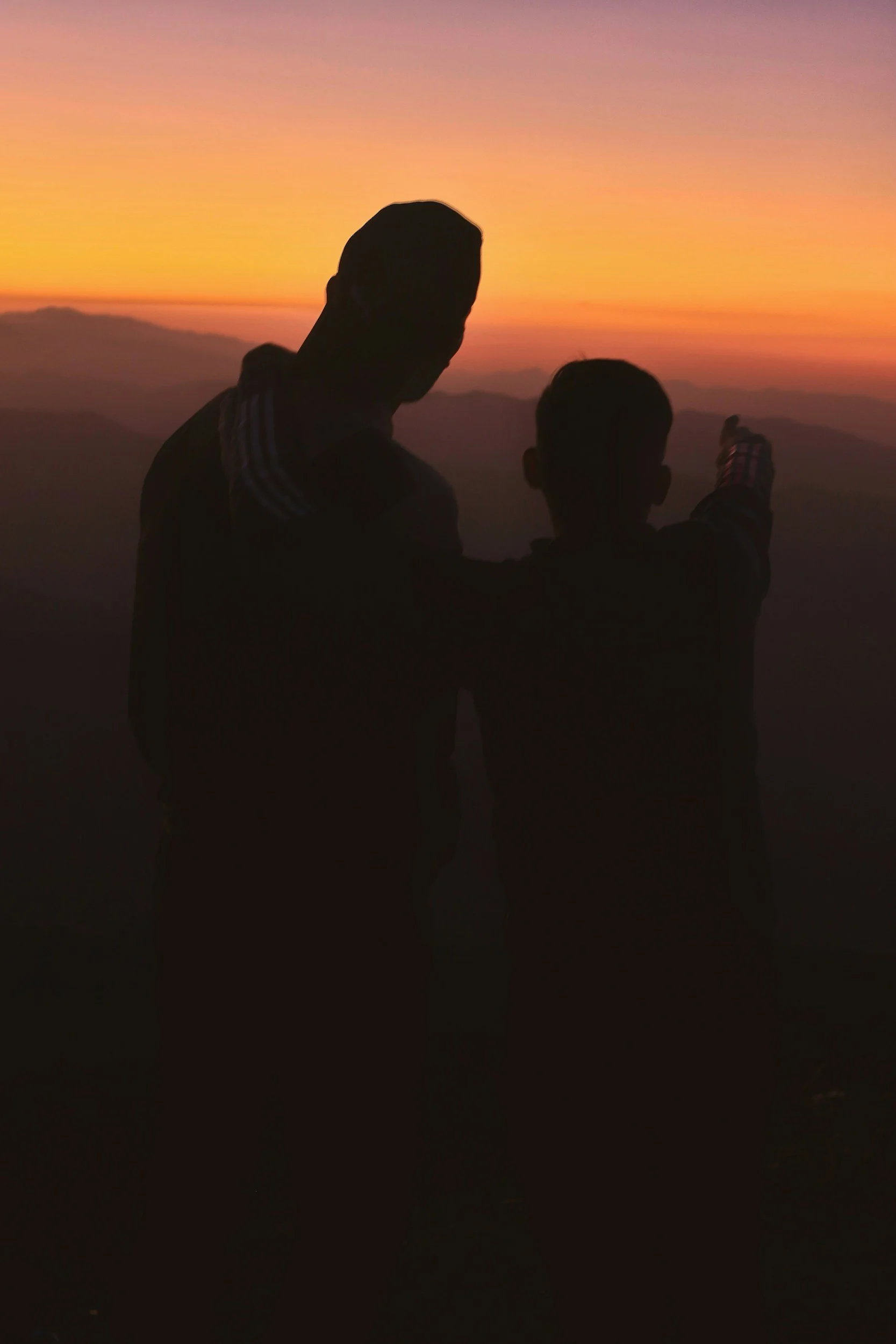 Silhouettes of two people standing on a mountain at sunset, one person pointing towards the horizon.