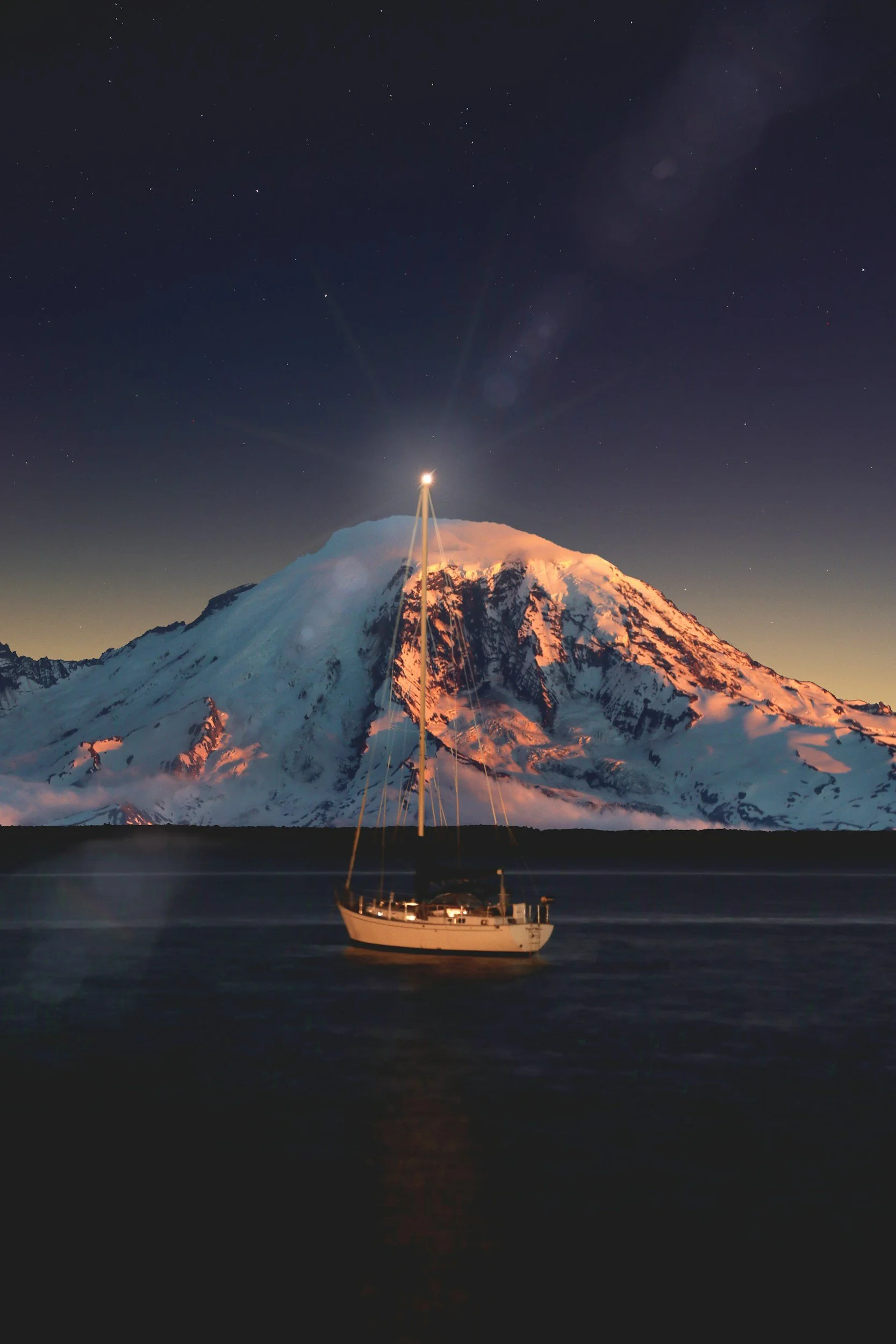 A sailboat on calm water in front of a snow-capped mountain at dusk, with stars visible in the sky.