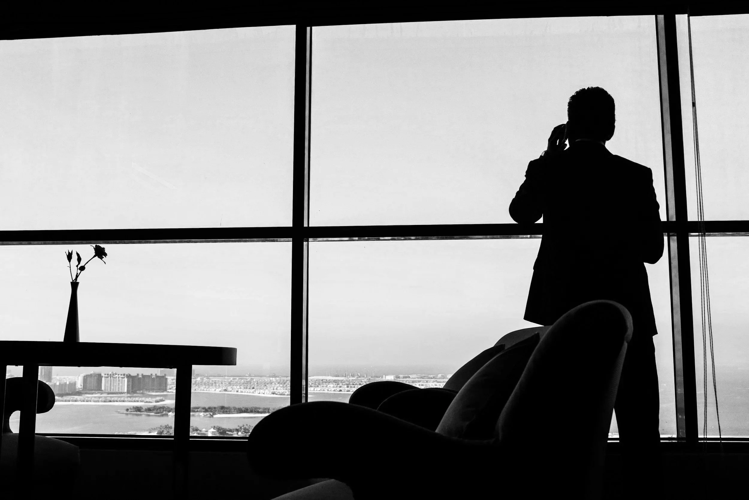 Silhouette of a man talking on a phone by a large window with a city view, a small table with a vase and flowers in the foreground.