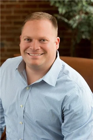 A smiling man in a light blue dress shirt sitting indoors with a brick wall and green plants in the background.