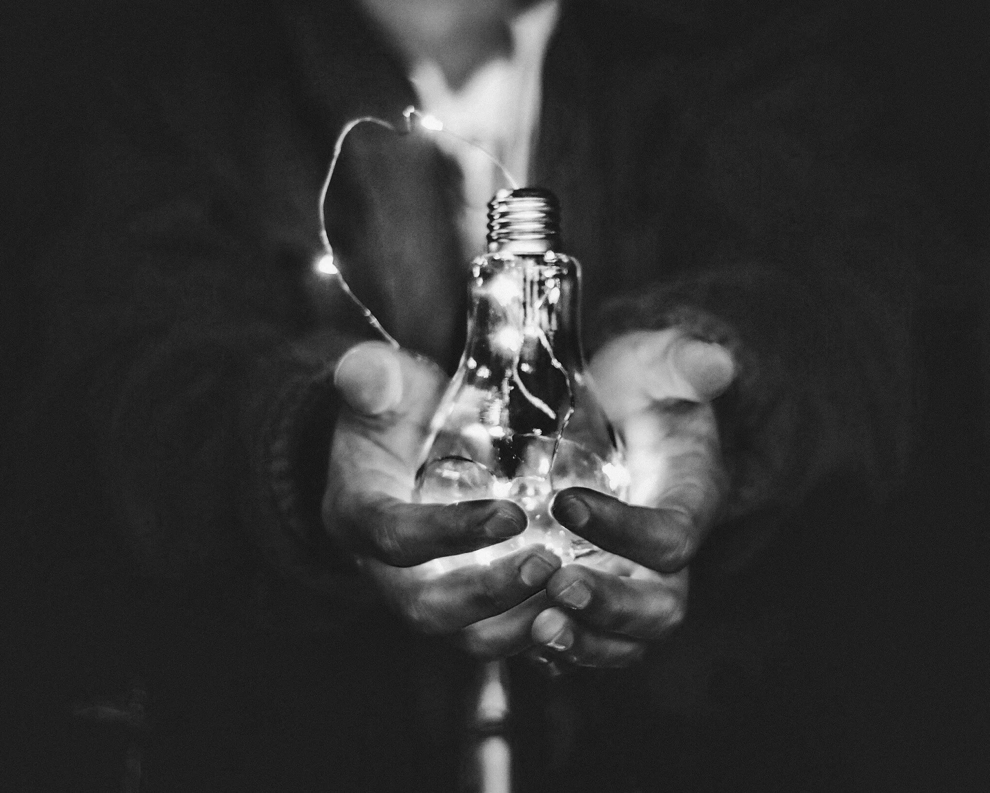 Black and white photo of hands holding a clear glass light bulb with a faint glow inside, surrounded by dark background.