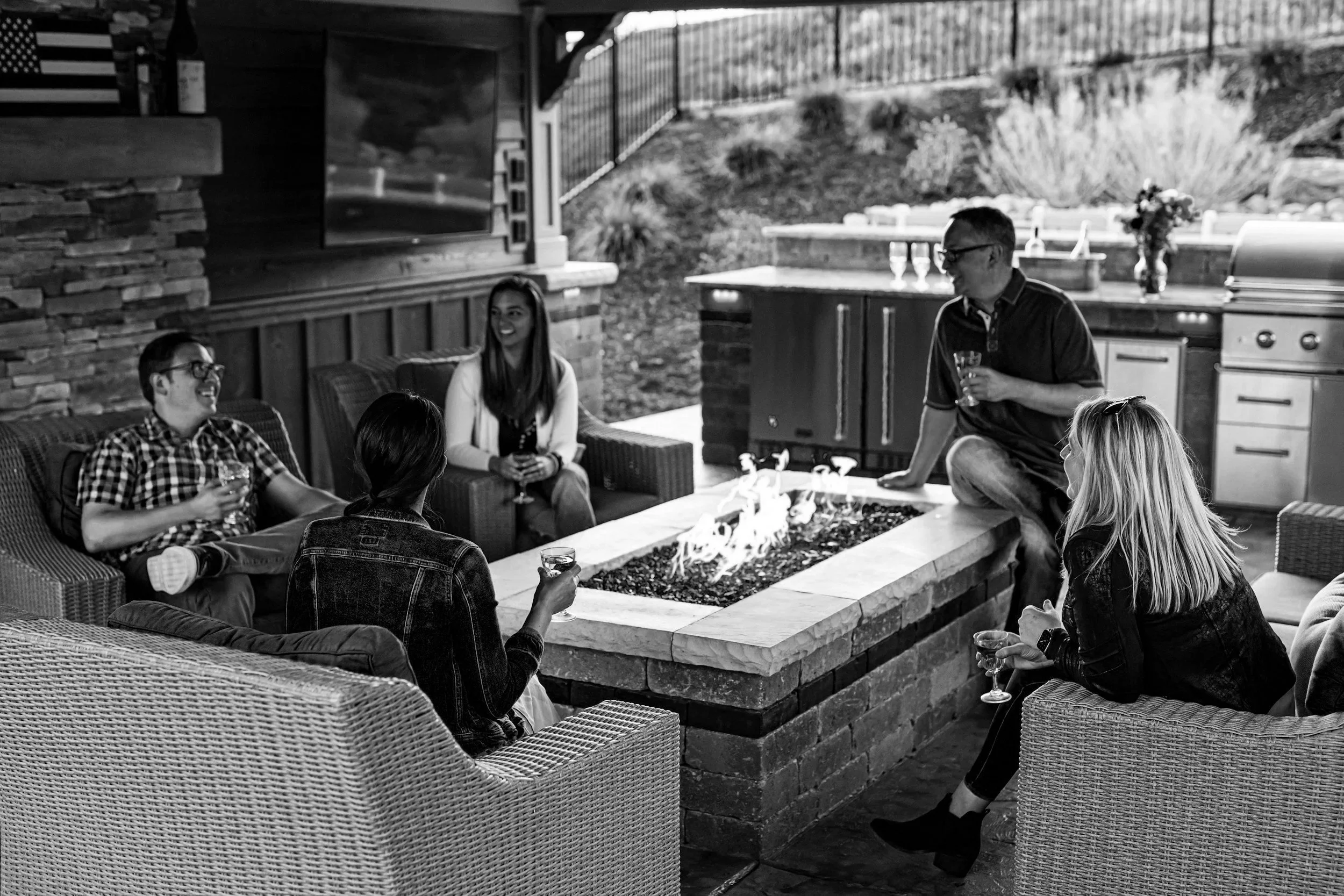 A group of people sitting around a fire pit on a patio, talking and drinking, with an outdoor kitchen in the background.
