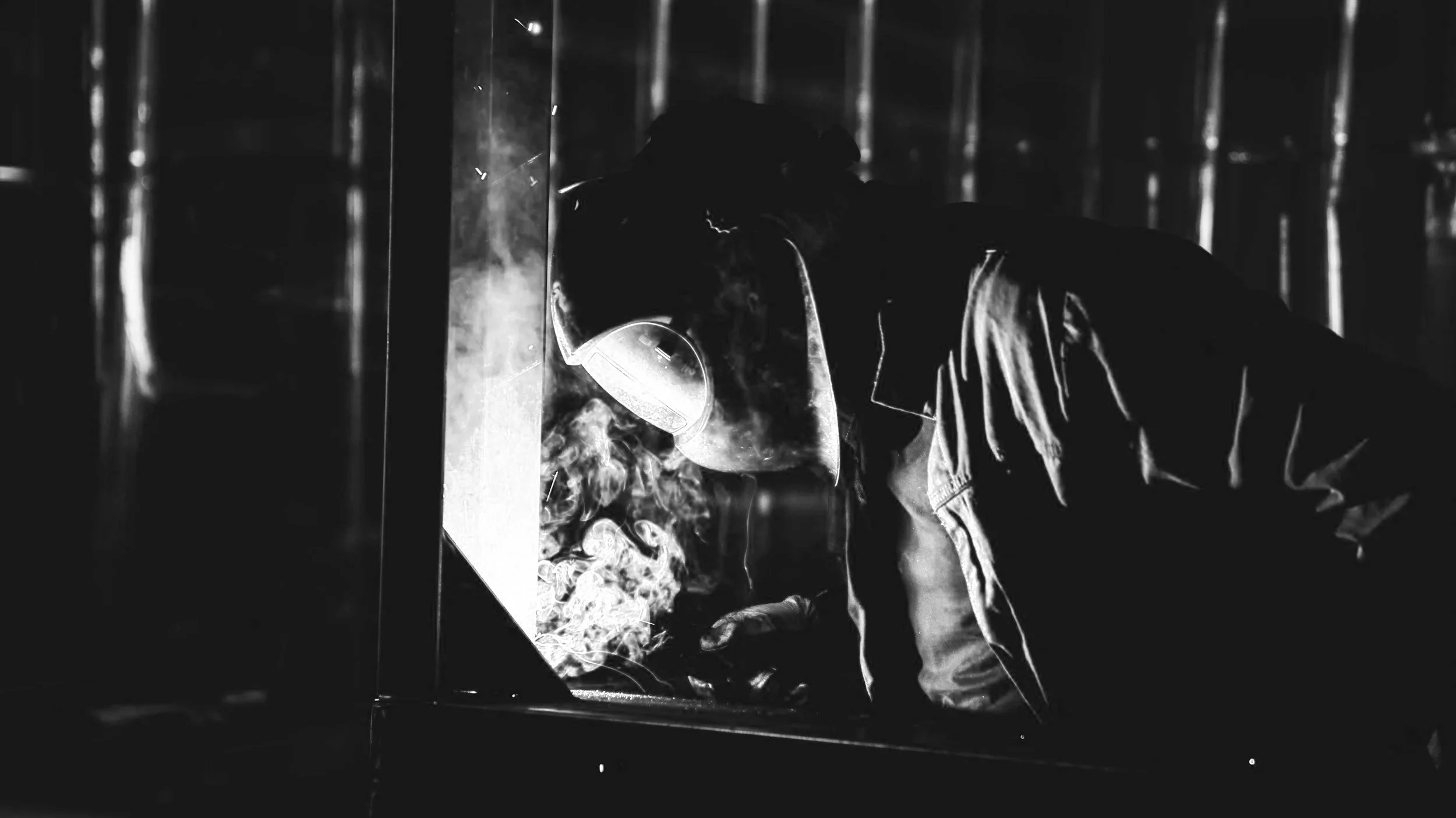 A welder wearing a protective mask and gloves welding metal, with sparks and smoke visible in a dark industrial setting.